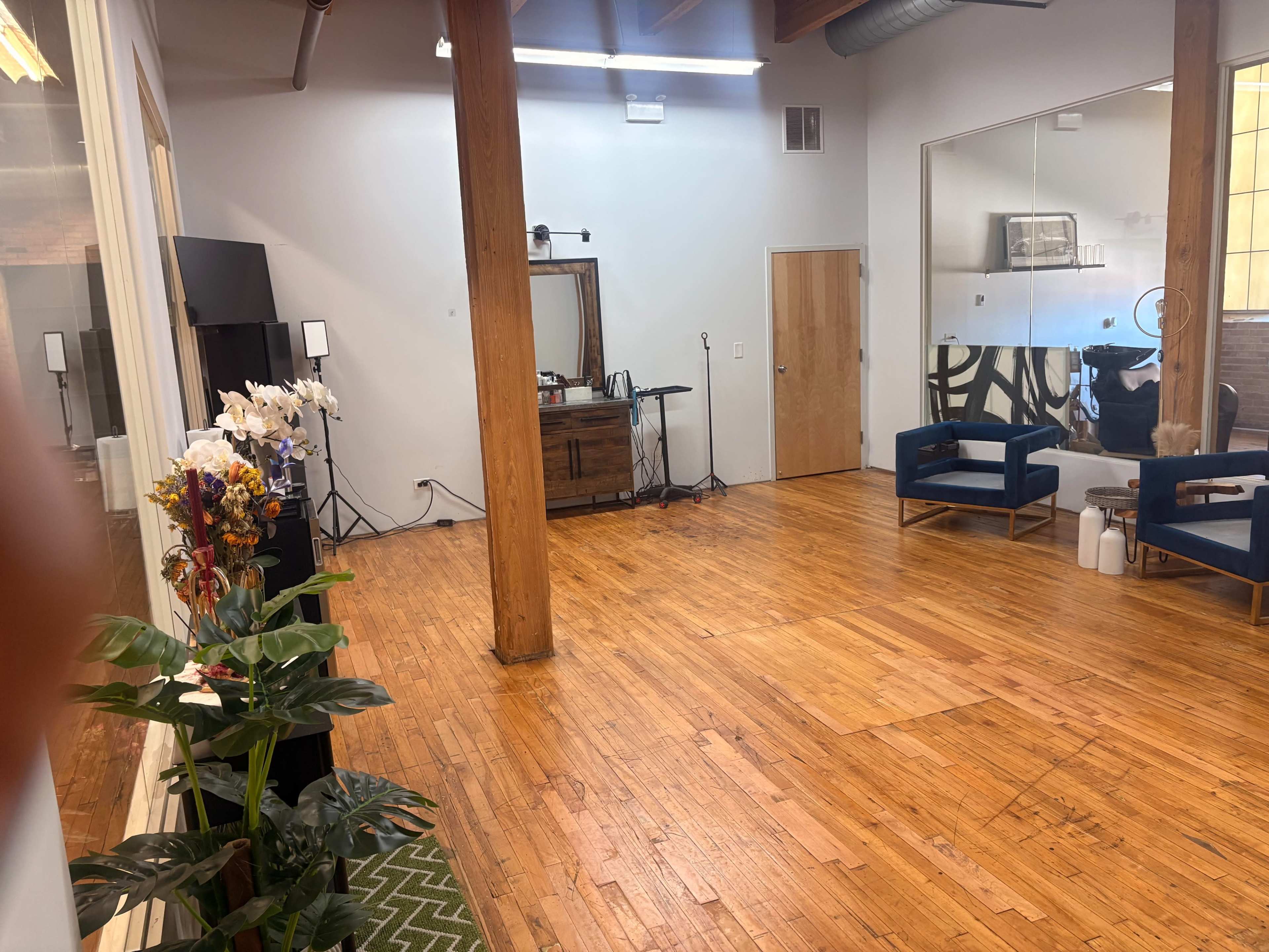 The image shows an empty, spacious room with wooden floors, large windows, and a seating area with two blue chairs and a console table along one wall.