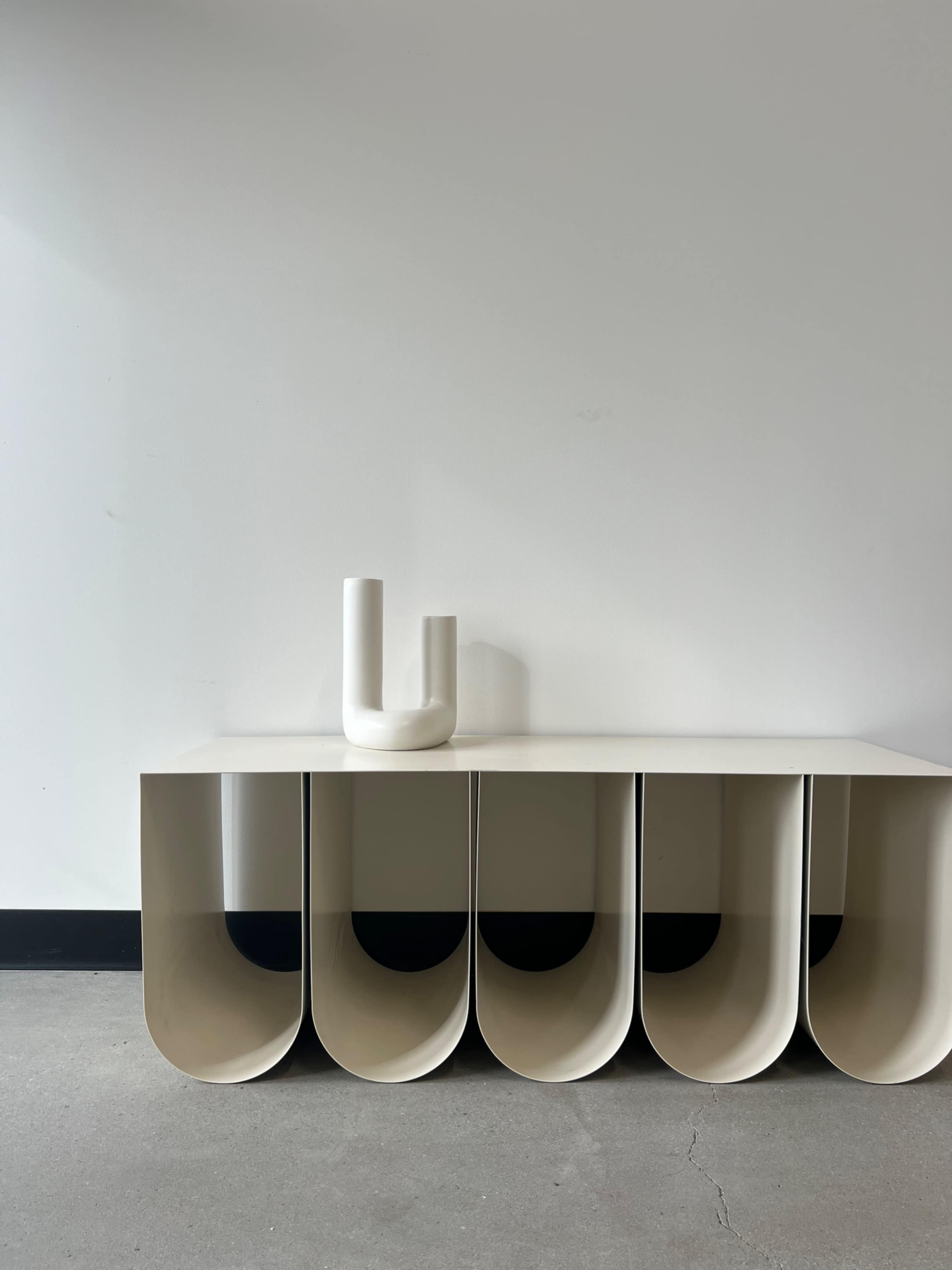 A minimalist white console table with curved compartments sits against a plain wall, topped by a simple white vase.