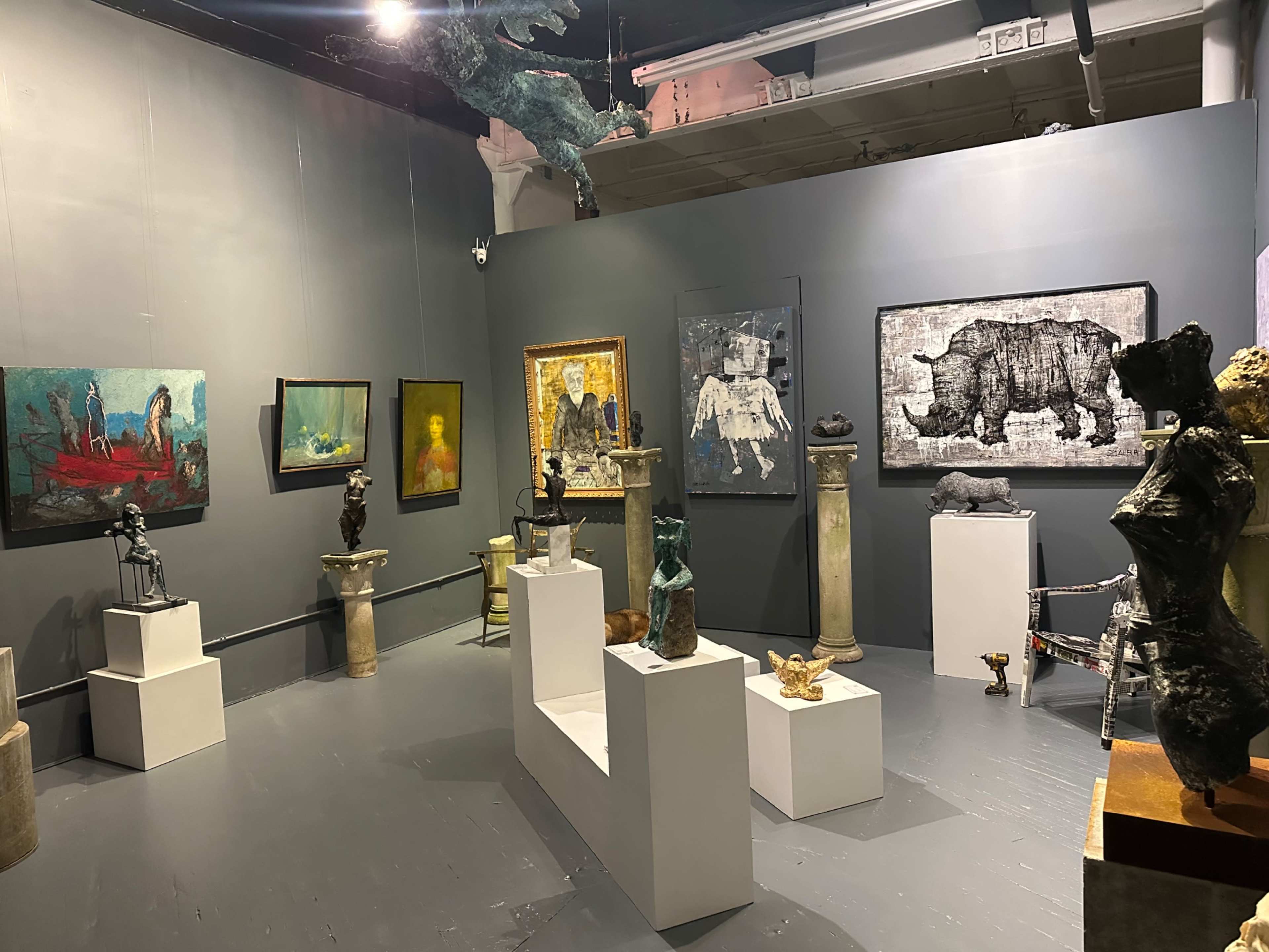 The image depicts an art gallery displaying various sculptures and paintings on gray walls, with artworks arranged on pedestals and hung at eye level.