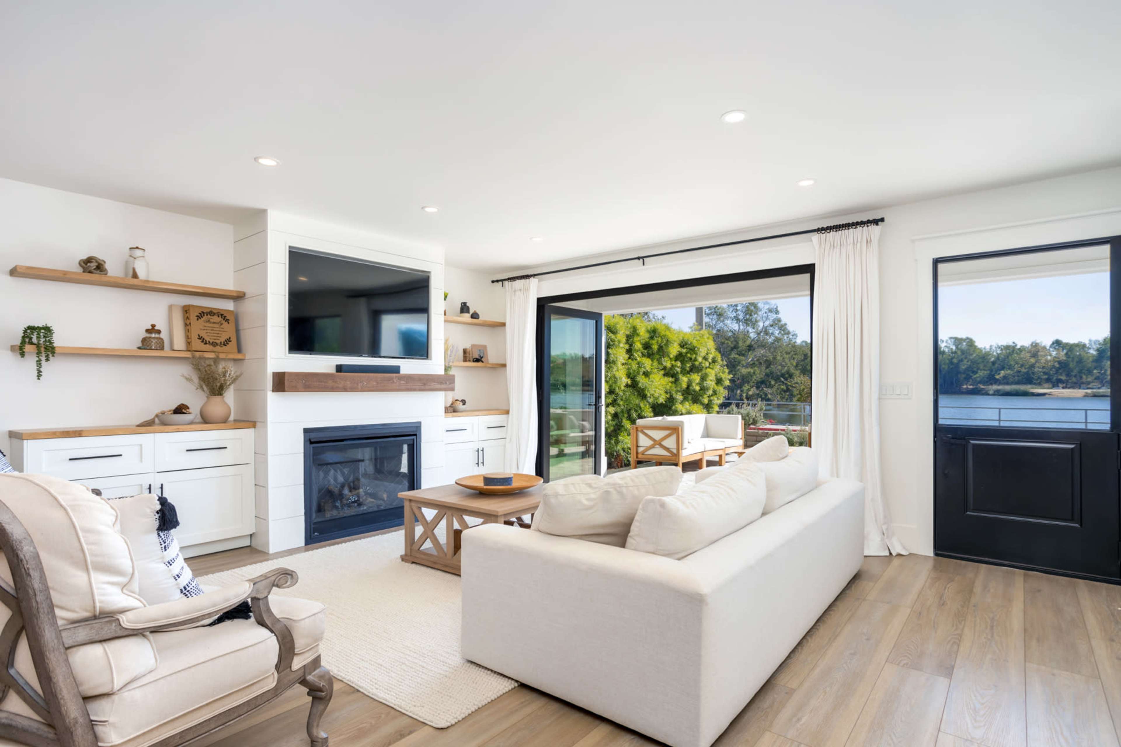 A modern living room features a white sofa, a wooden coffee table, and a television mounted on a light-colored wall, with large windows overlooking a scenic view.