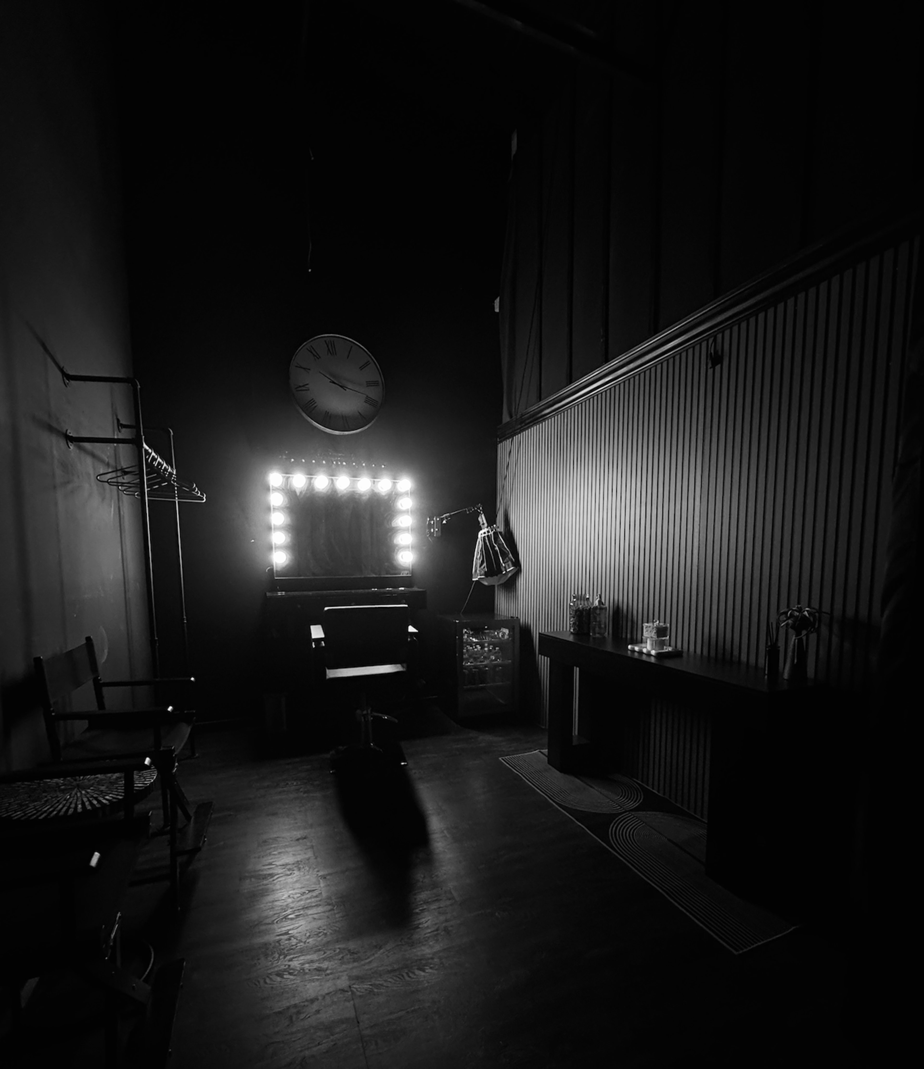 The image shows a dimly lit room with a makeup mirror surrounded by lights, a clock on the wall, and simple furniture including a chair and a table.