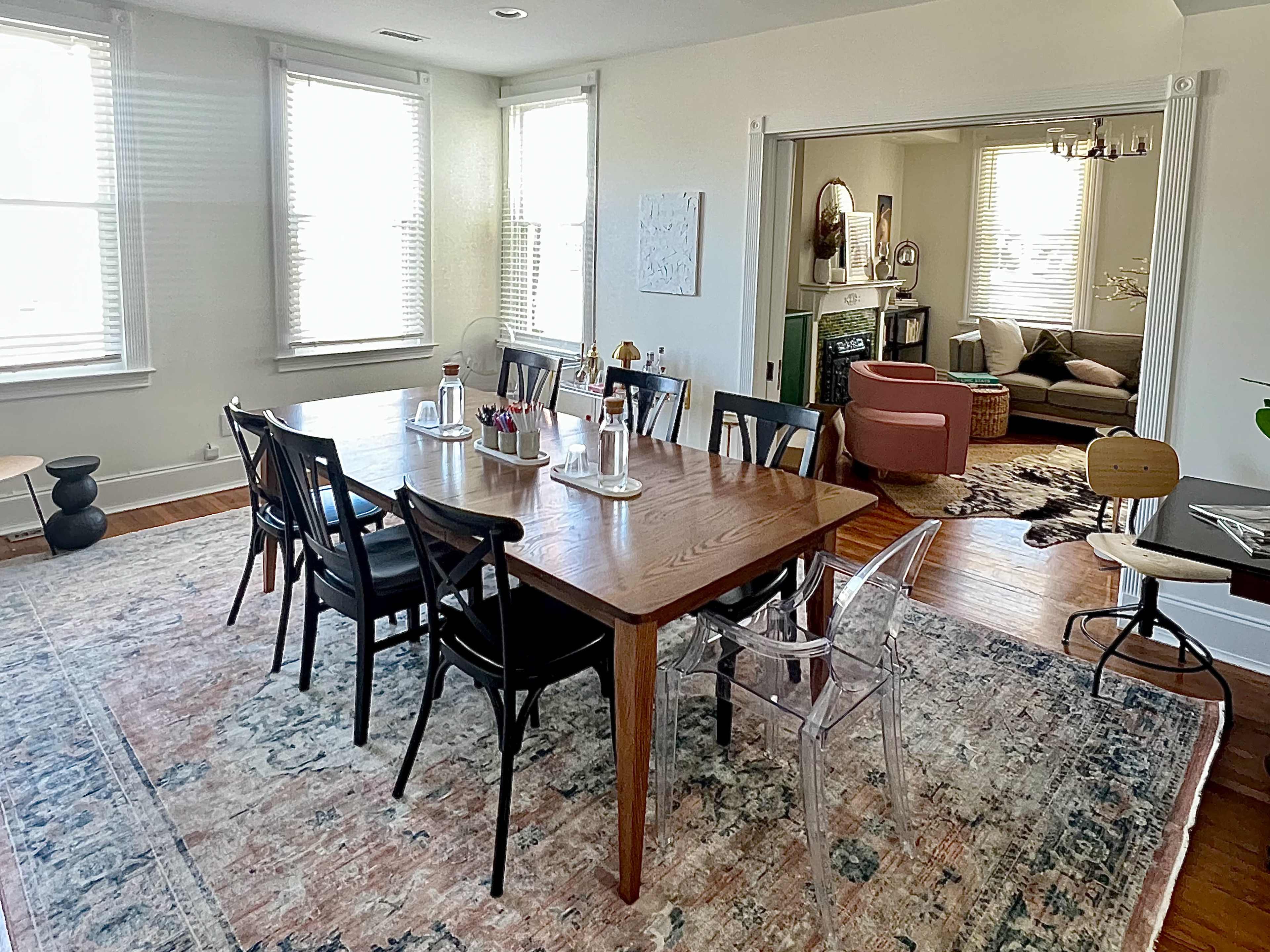 The image shows a dining area with a large wooden table surrounded by black chairs, adjacent to a living room with a couch and decorative elements.
