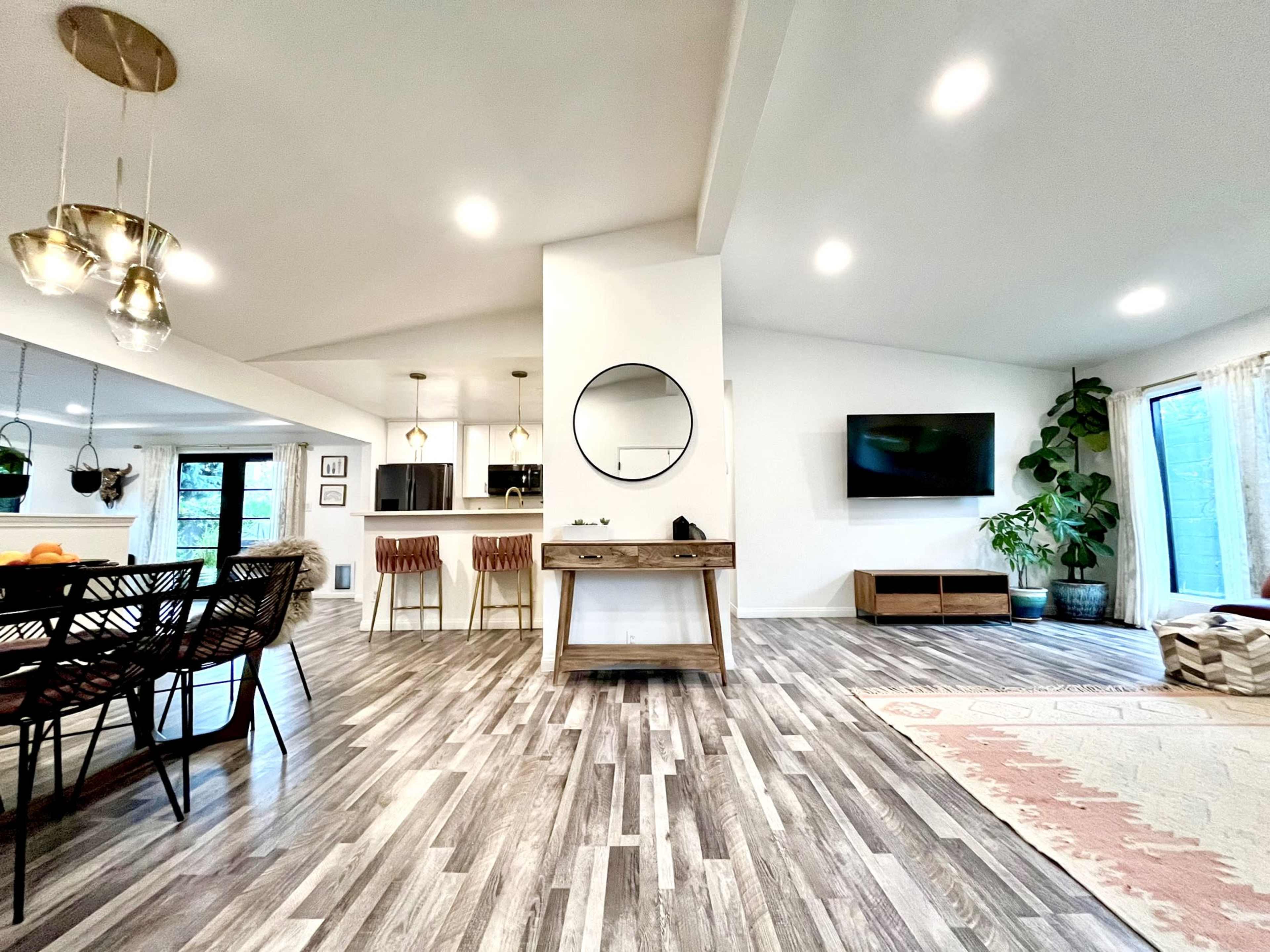 An open-concept living and dining area with wooden flooring, a circular mirror on the wall, and large windows allowing natural light to enter.