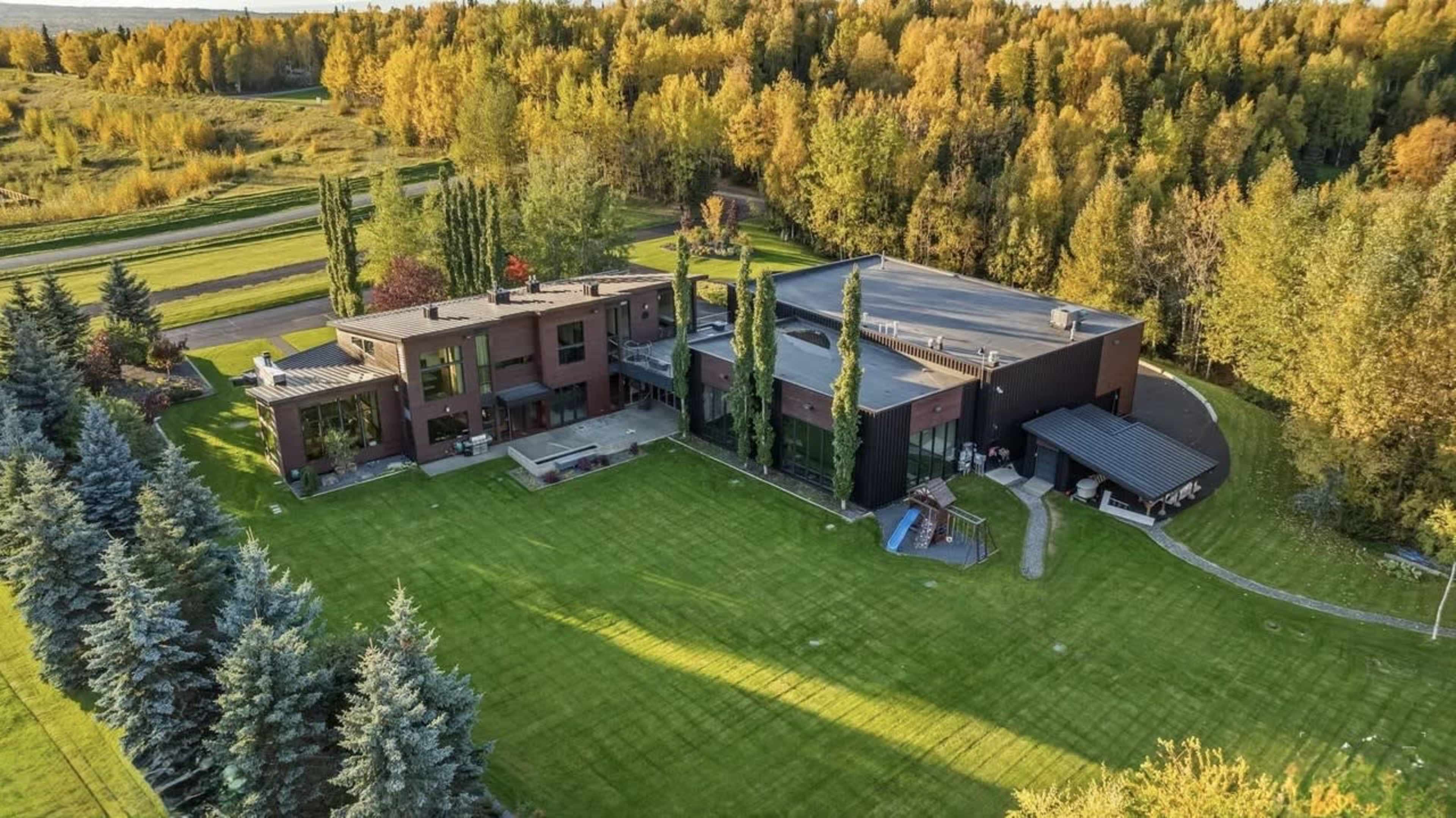 The image shows a large modern house surrounded by well-manicured lawns and lined with trees, set against a backdrop of autumn foliage.