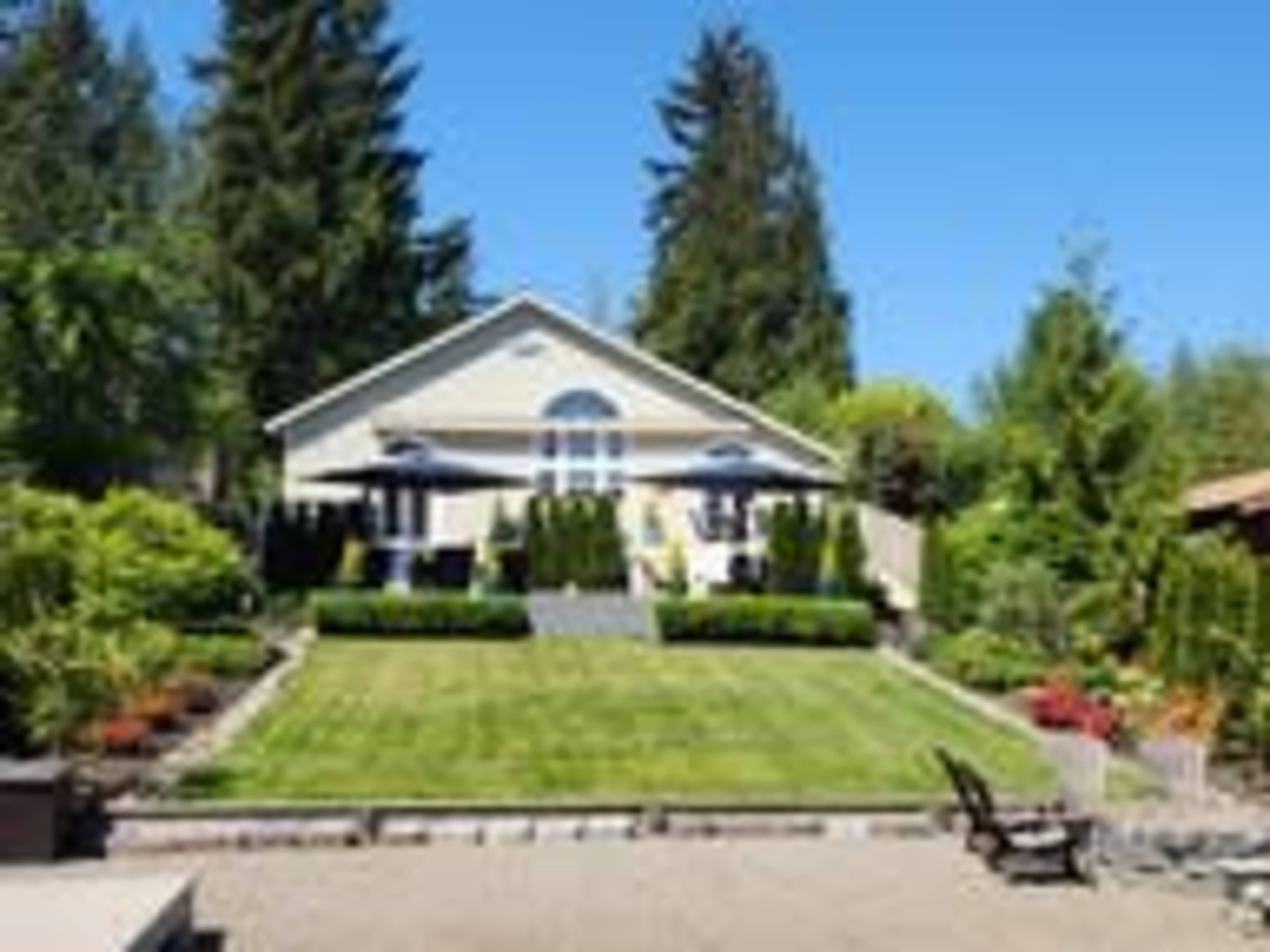 A well-maintained house is set back with a manicured lawn and surrounded by trees and shrubs.