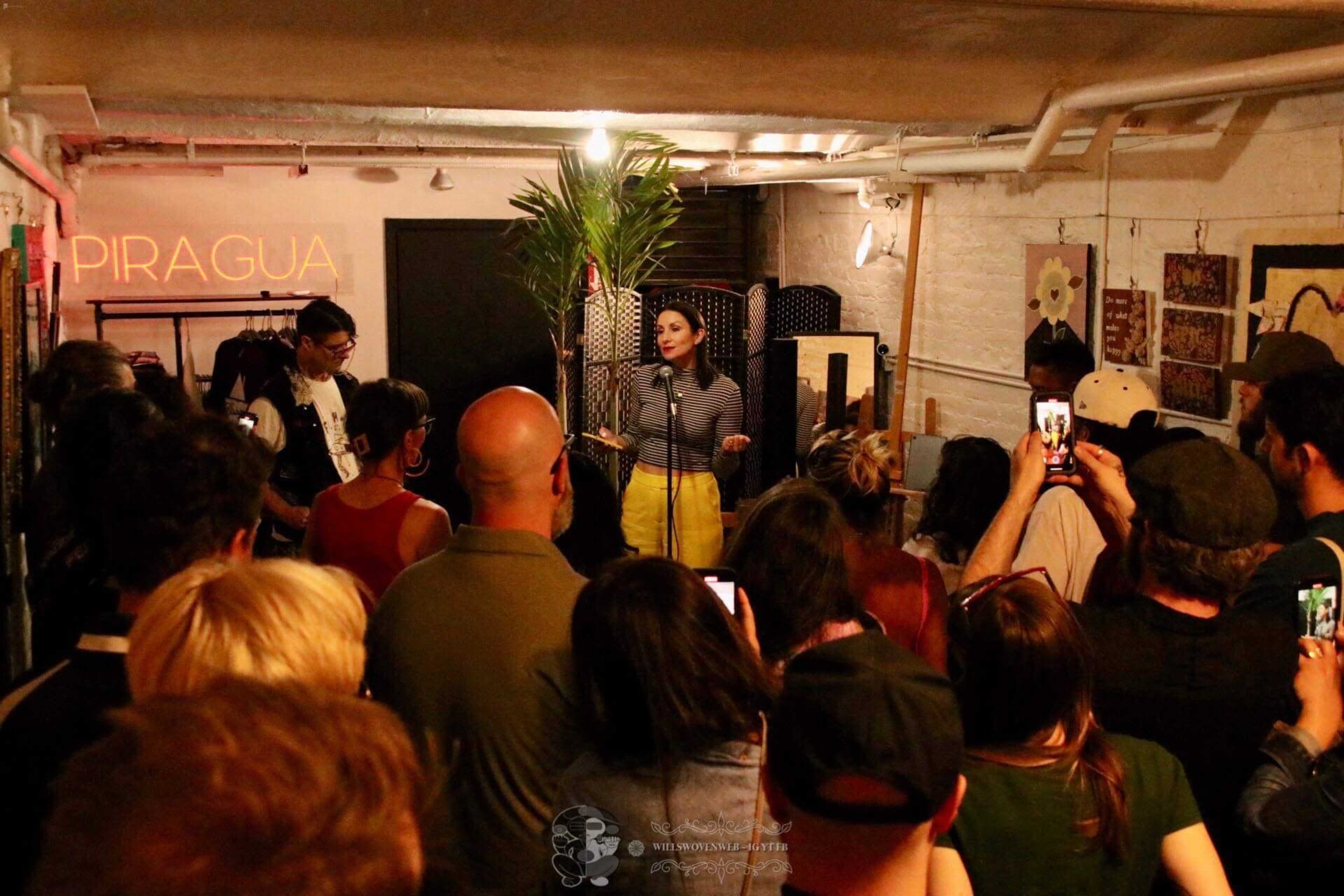 A singer performs on stage in front of an audience in a dimly lit venue filled with clothing and decor.