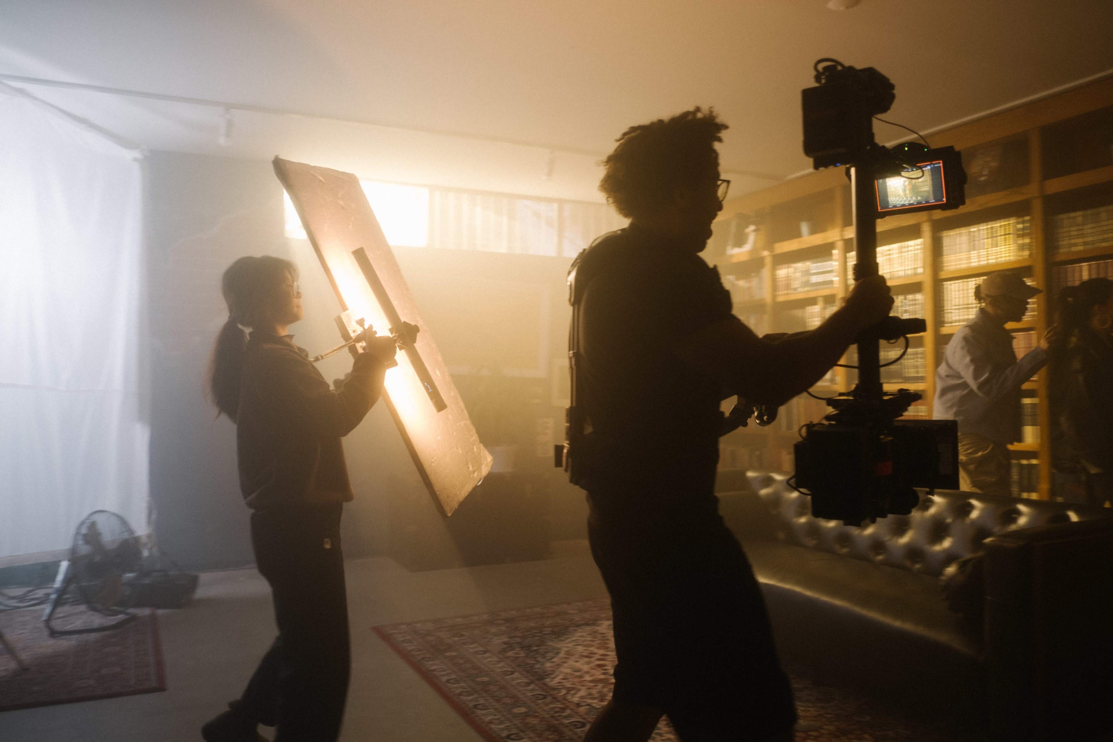 A crew member holds a reflector while another operates a camera in a dimly lit room filled with shelves of books.
