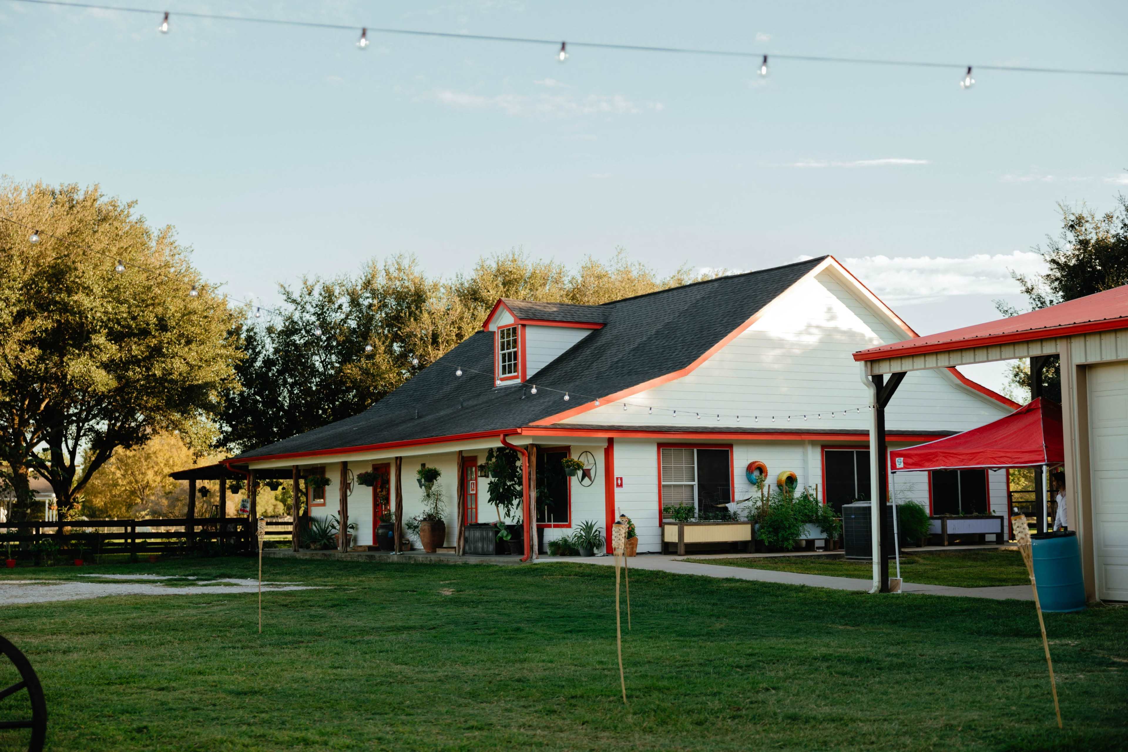 A white house with red accents sits in a grassy area, surrounded by trees and string lights.