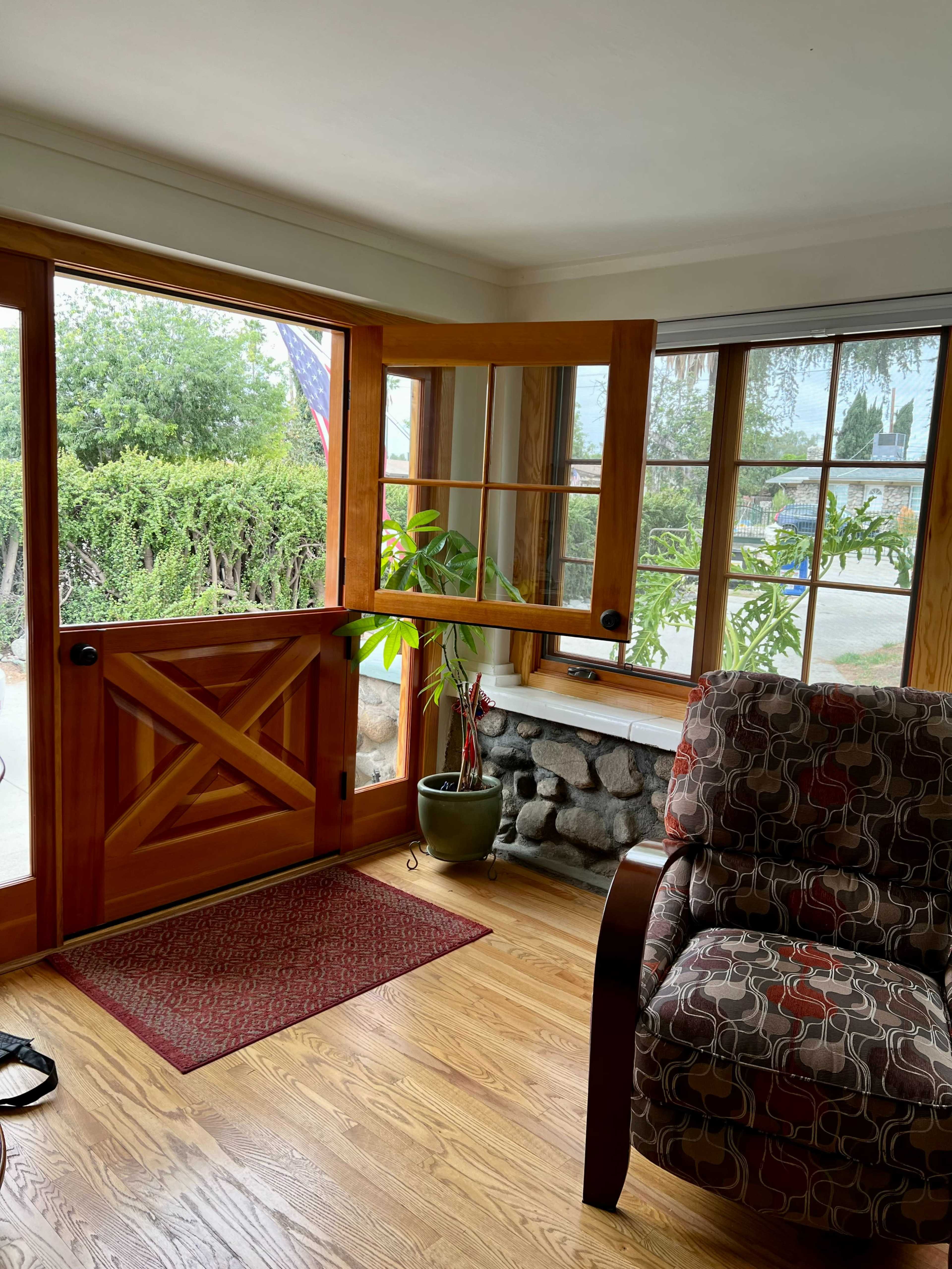 The image shows a bright interior corner of a room featuring a wooden door with a large window, a potted plant, a patterned chair, and a stone fireplace.