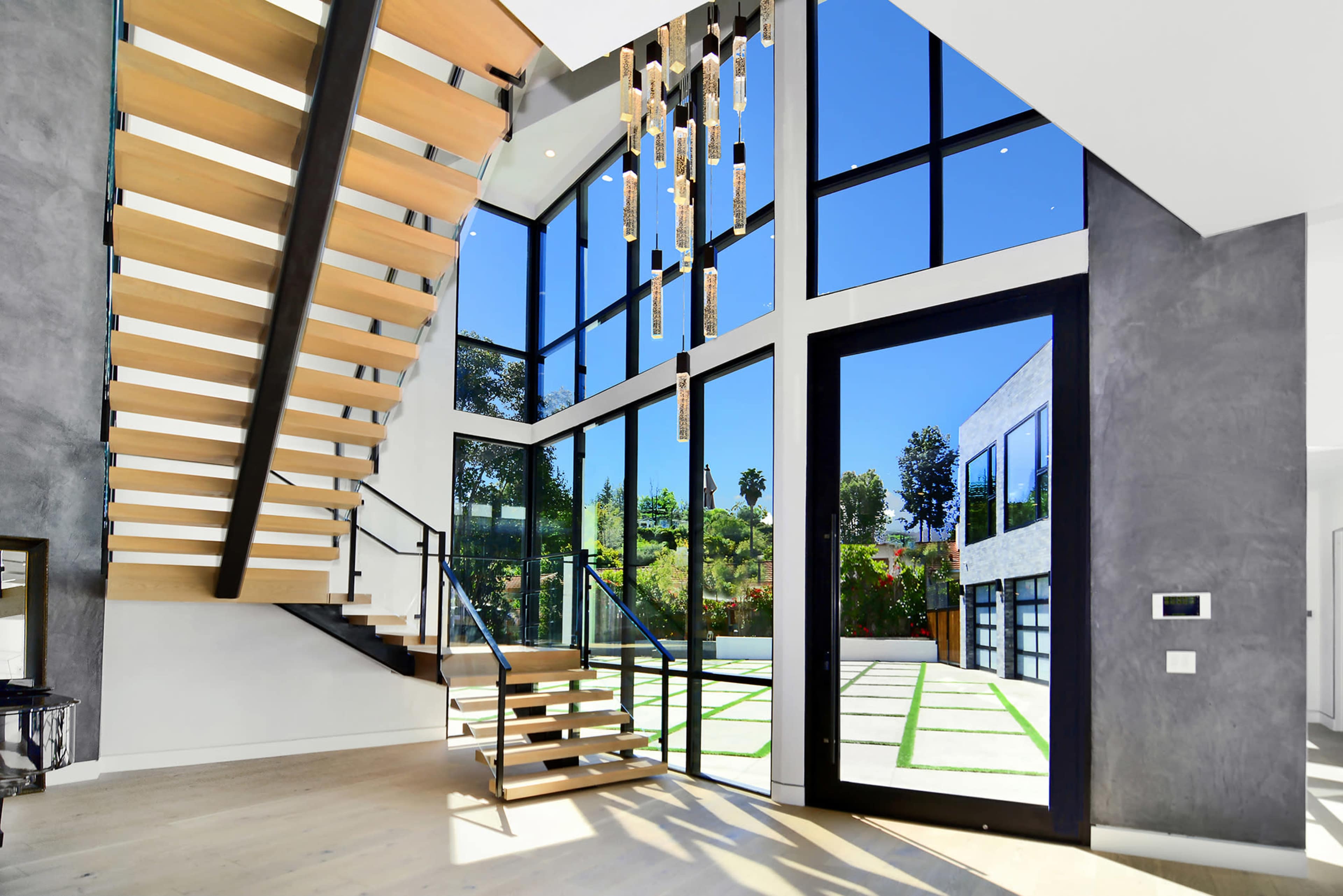 A modern interior space with a prominent wooden staircase, large glass windows, and a view of greenery outside.