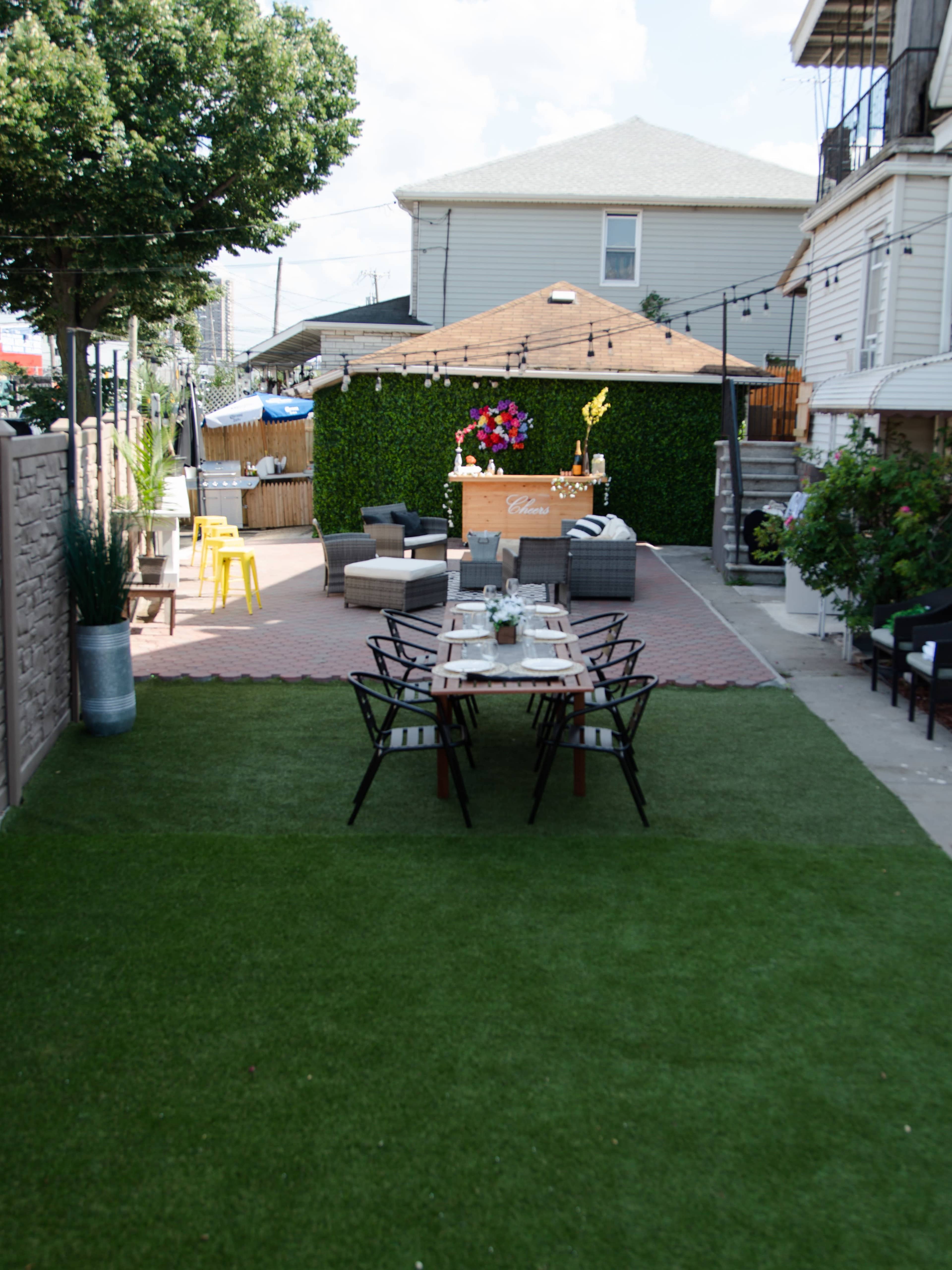 A backyard space with green turf, a dining table with chairs, and a lounge area surrounded by shrubs and wooden structures.