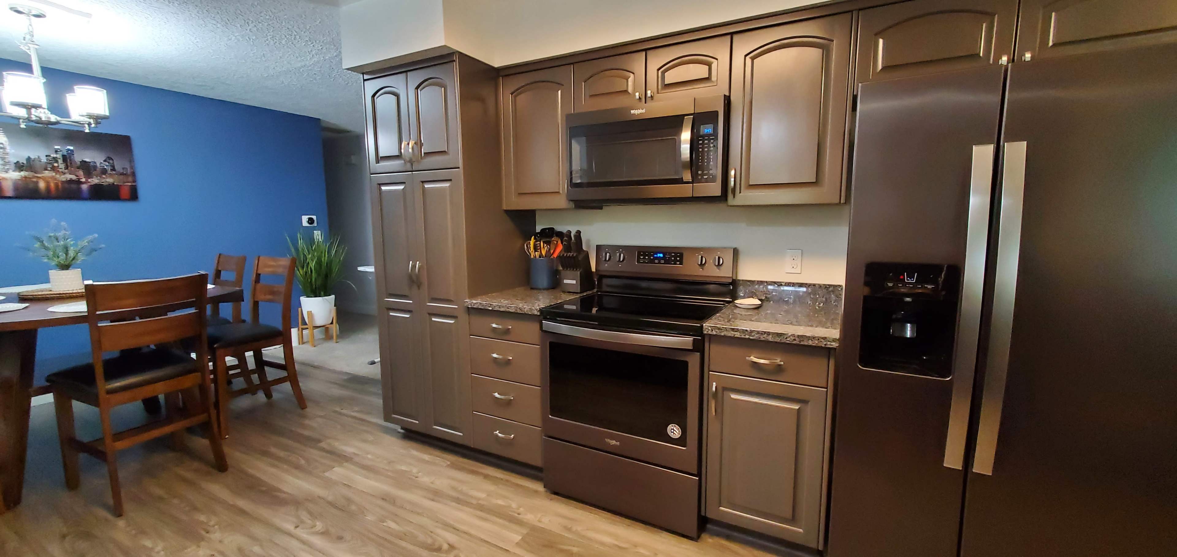 The image shows a modern kitchen with dark cabinets, a stainless steel refrigerator, a microwave over the stove, and a dining area in the background featuring a wooden table and chairs.