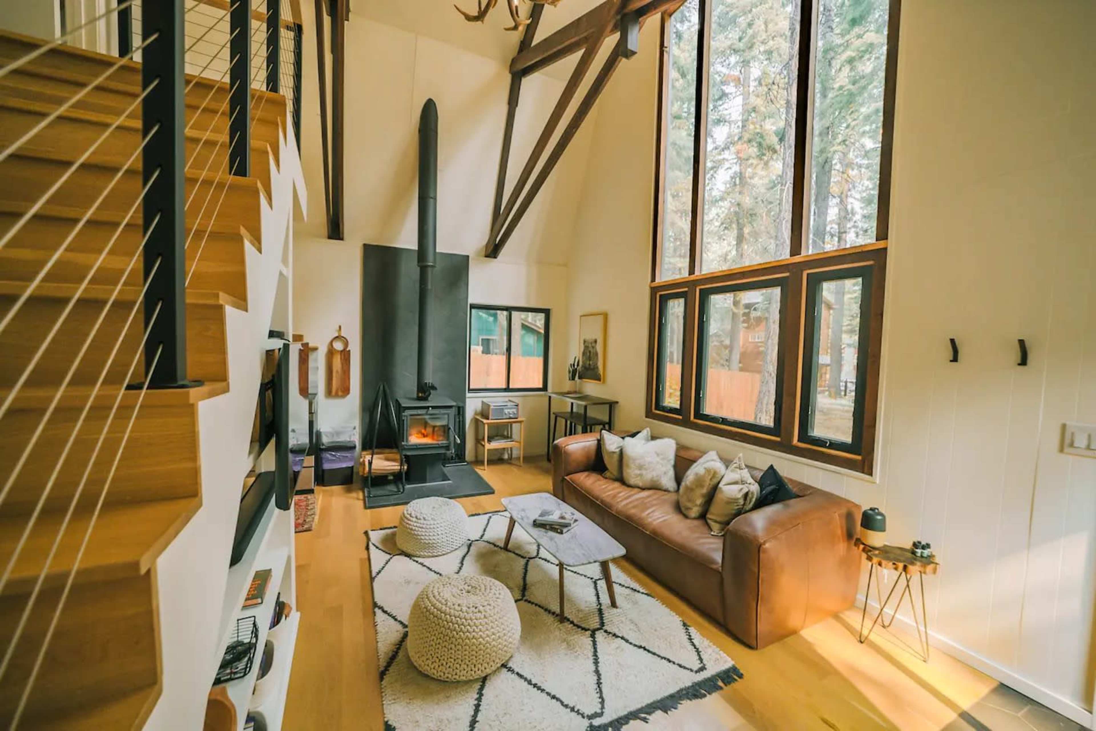 A modern living room with tall windows, a fireplace, a brown leather couch, and a staircase leading to a loft area.