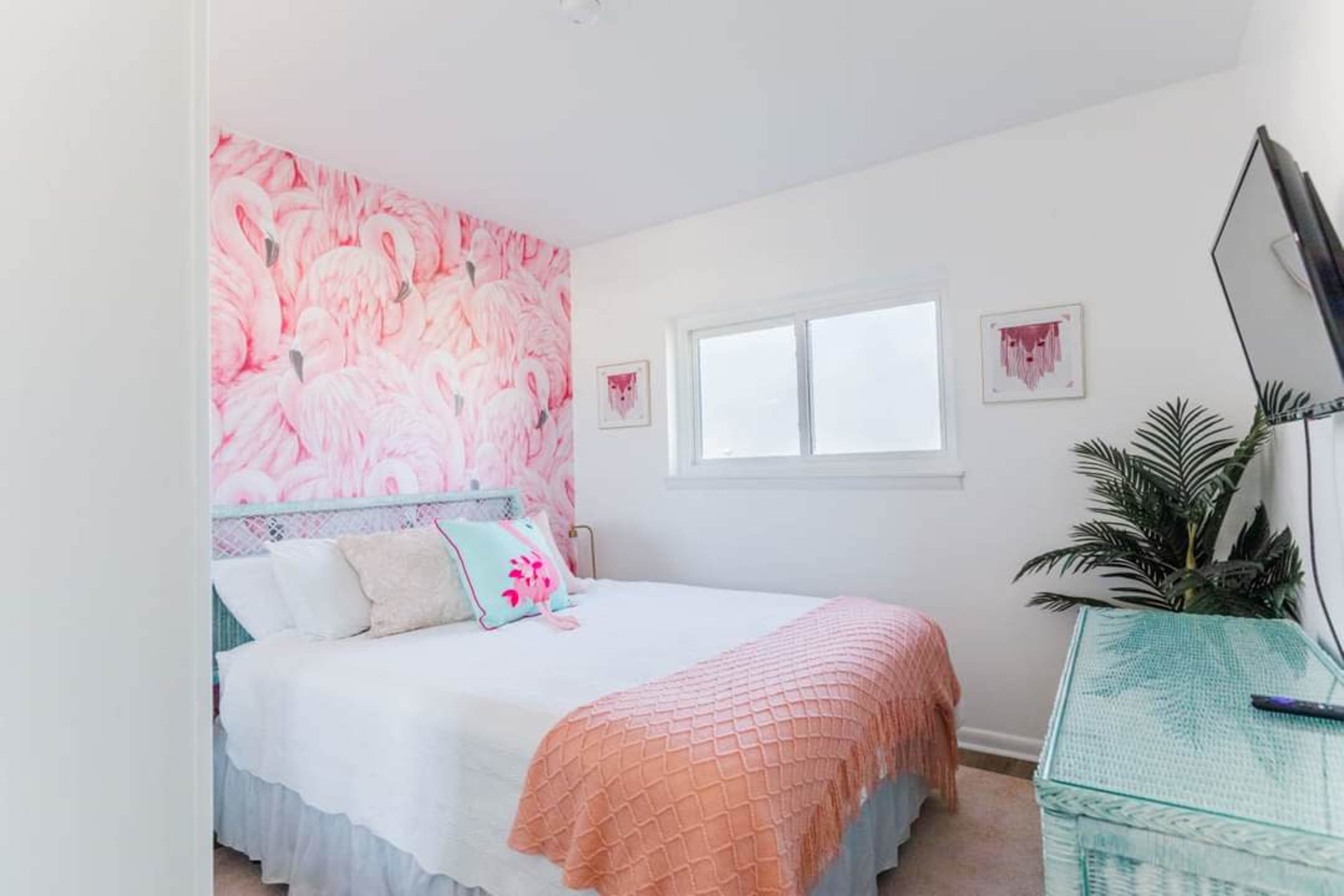 A bedroom features a pink flamingo wallpaper, a white bed with a textured coral throw, and a window beside a decorative plant.