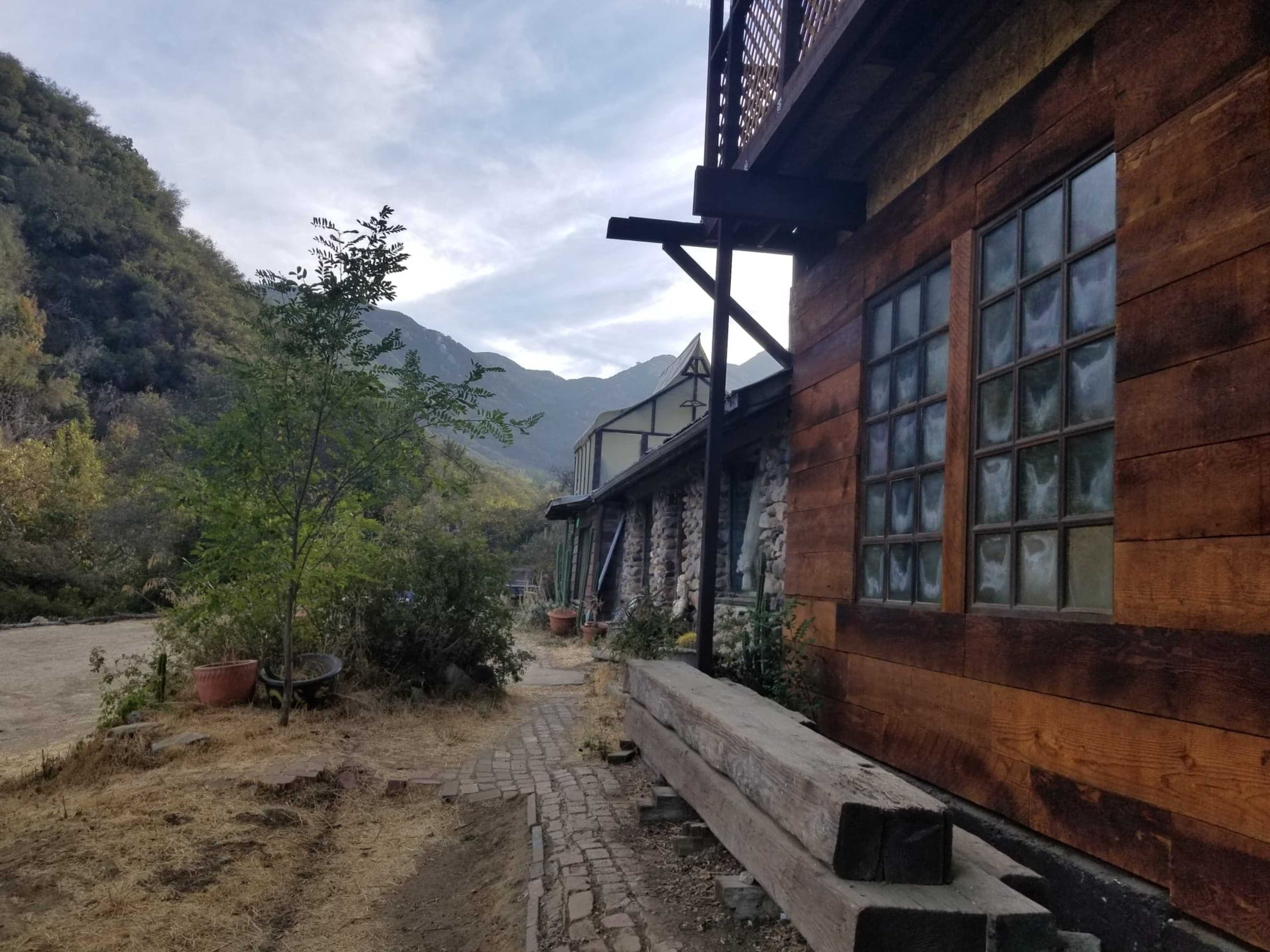 A rustic building made of wood and stone is situated alongside a dirt path, with mountains visible in the background.