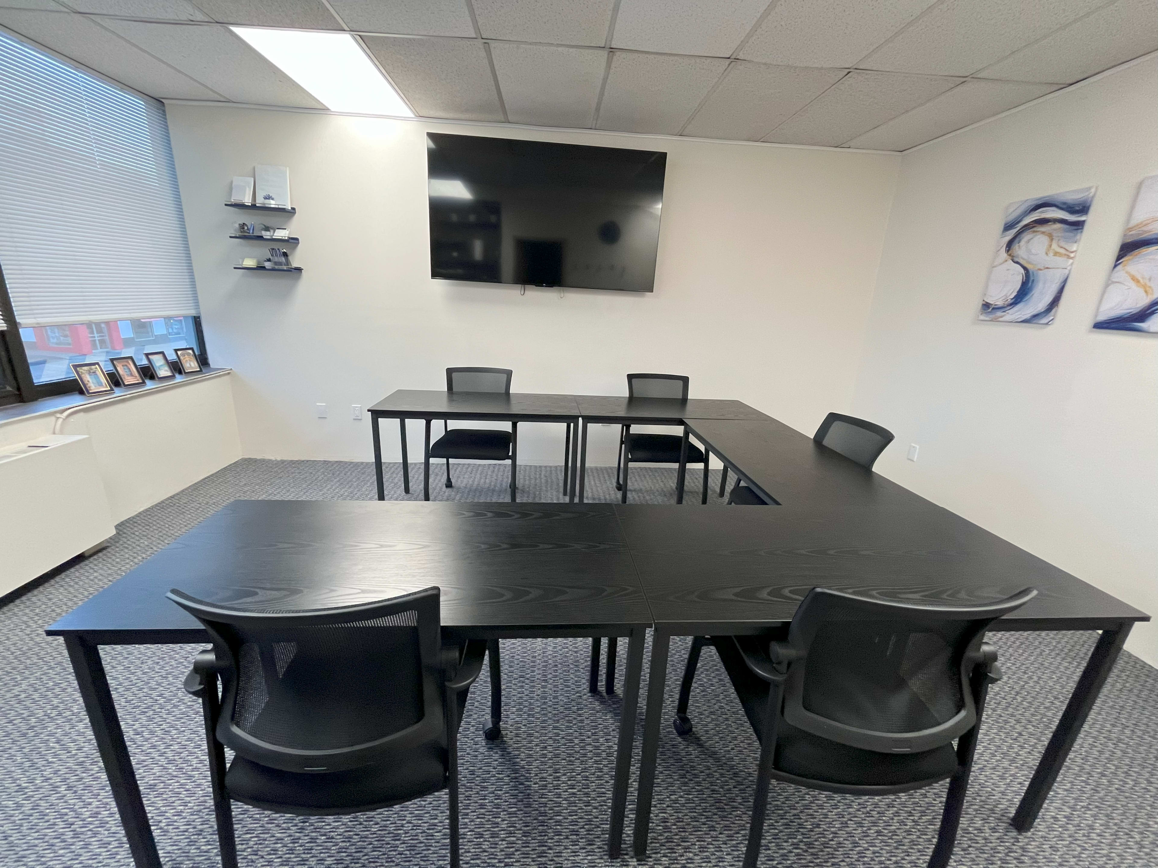 A small conference room features two rectangular black tables arranged in a T-shape, surrounded by black chairs, with a wall-mounted TV and framed photos on a shelf.