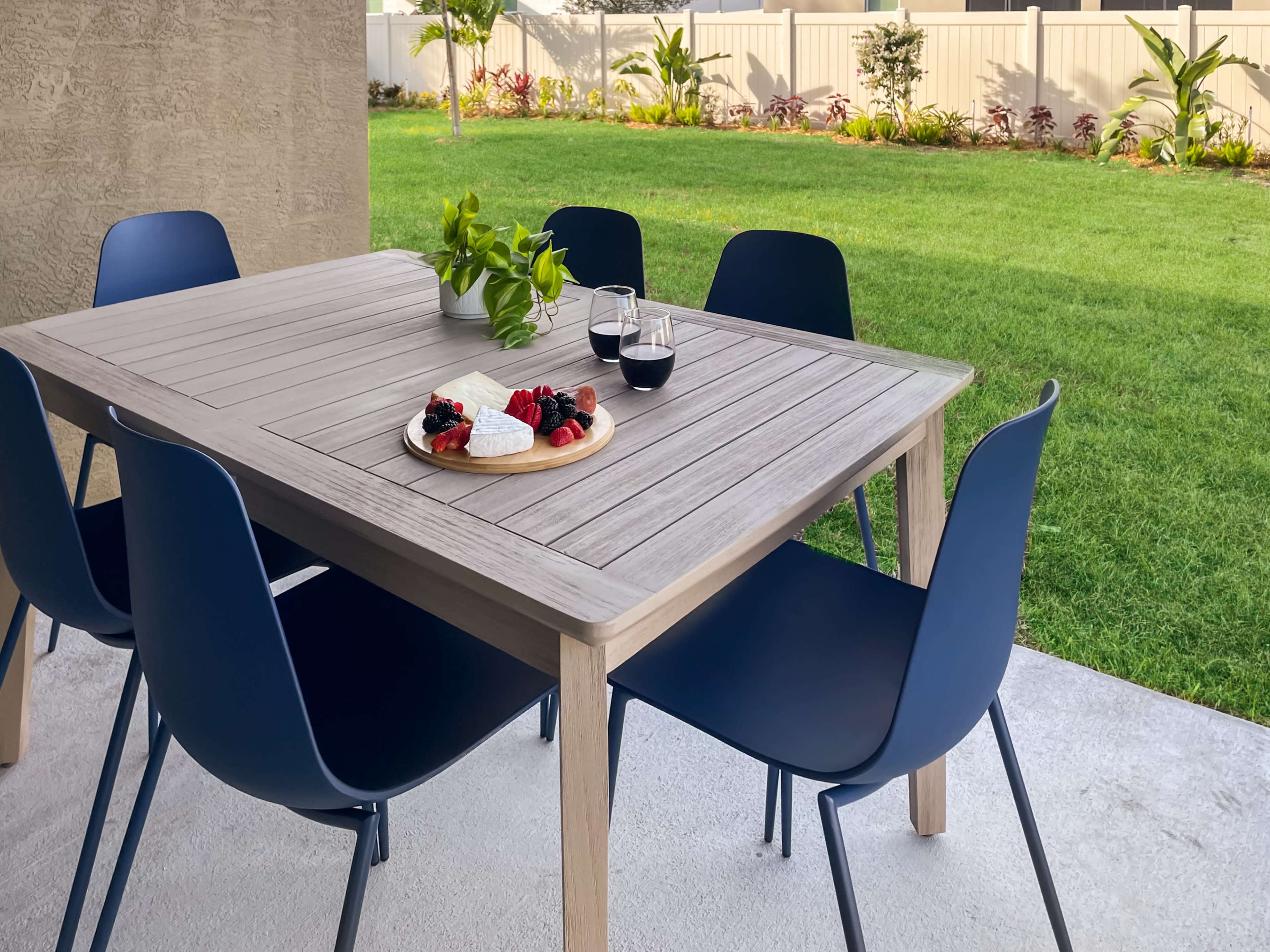 A wooden outdoor dining table is set on a patio, surrounded by six blue chairs, with a plate of fruit and two glasses of dark liquid on the table, and a grassy yard in the background.