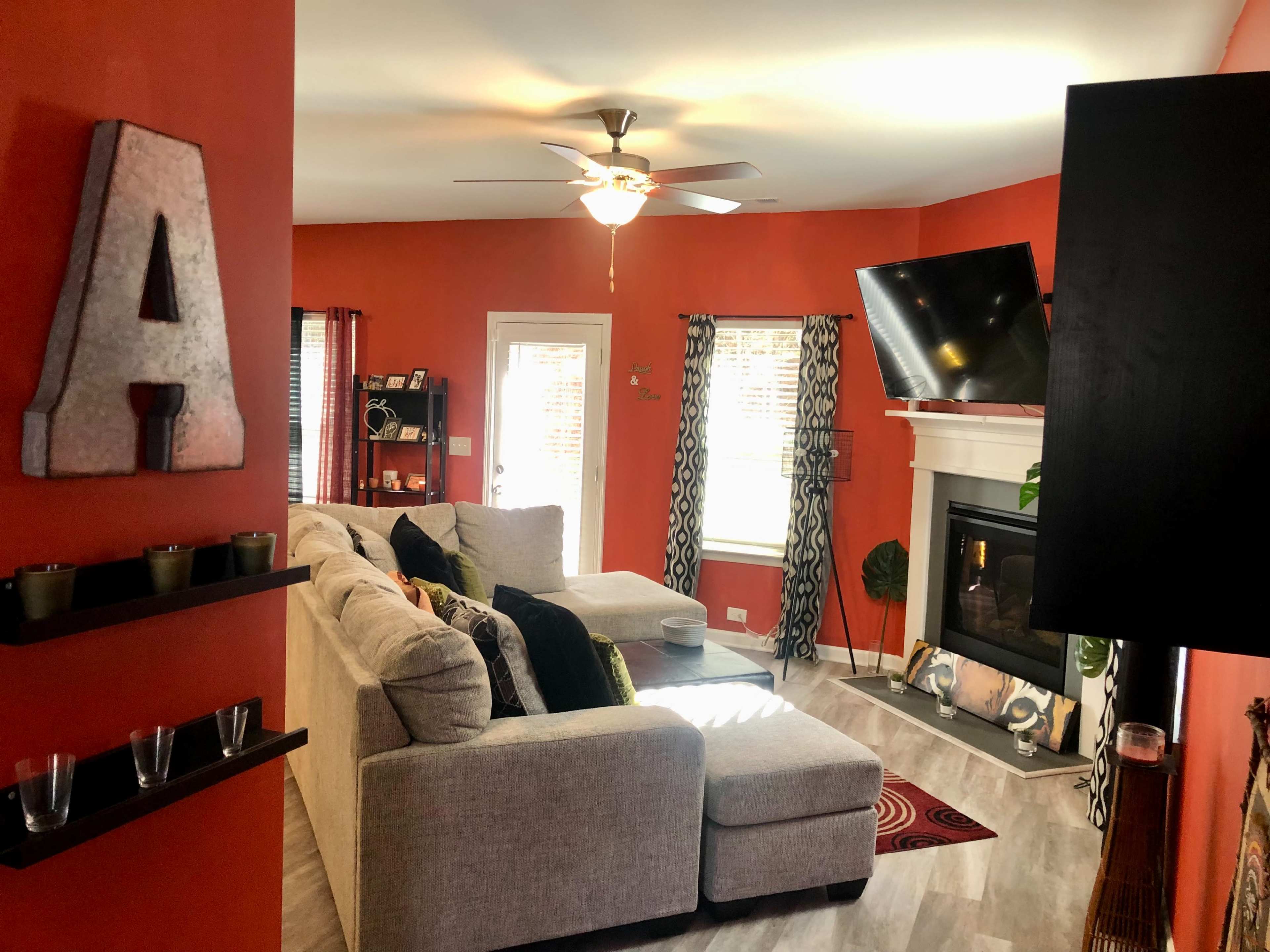 A cozy living room features a gray sectional sofa, a ceiling fan, and a television mounted above a fireplace, with orange walls and decorative accents.