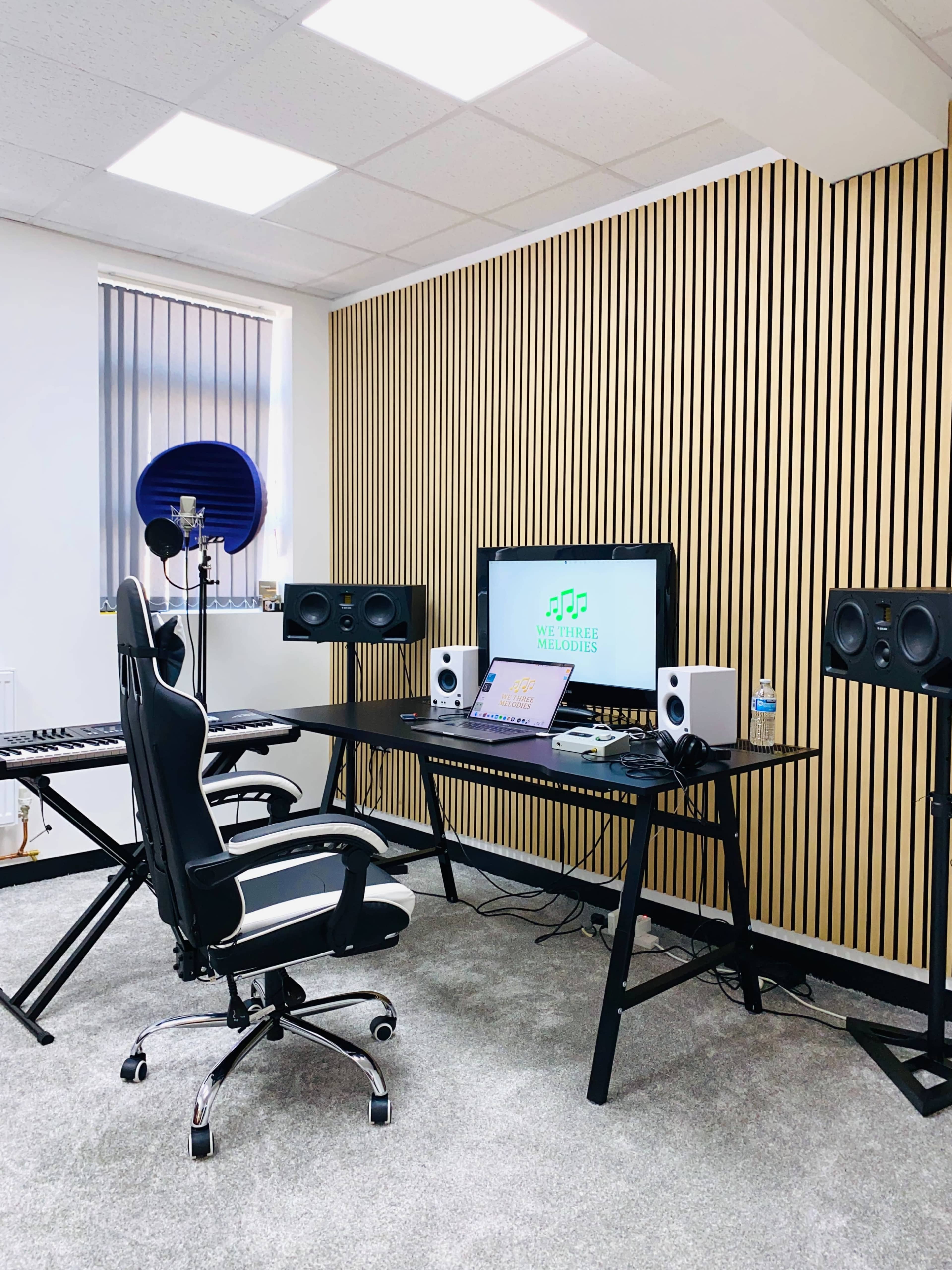 The image shows a music production studio featuring a desk with a computer, studio monitors, a keyboard, and a microphone against a wooden slatted wall.