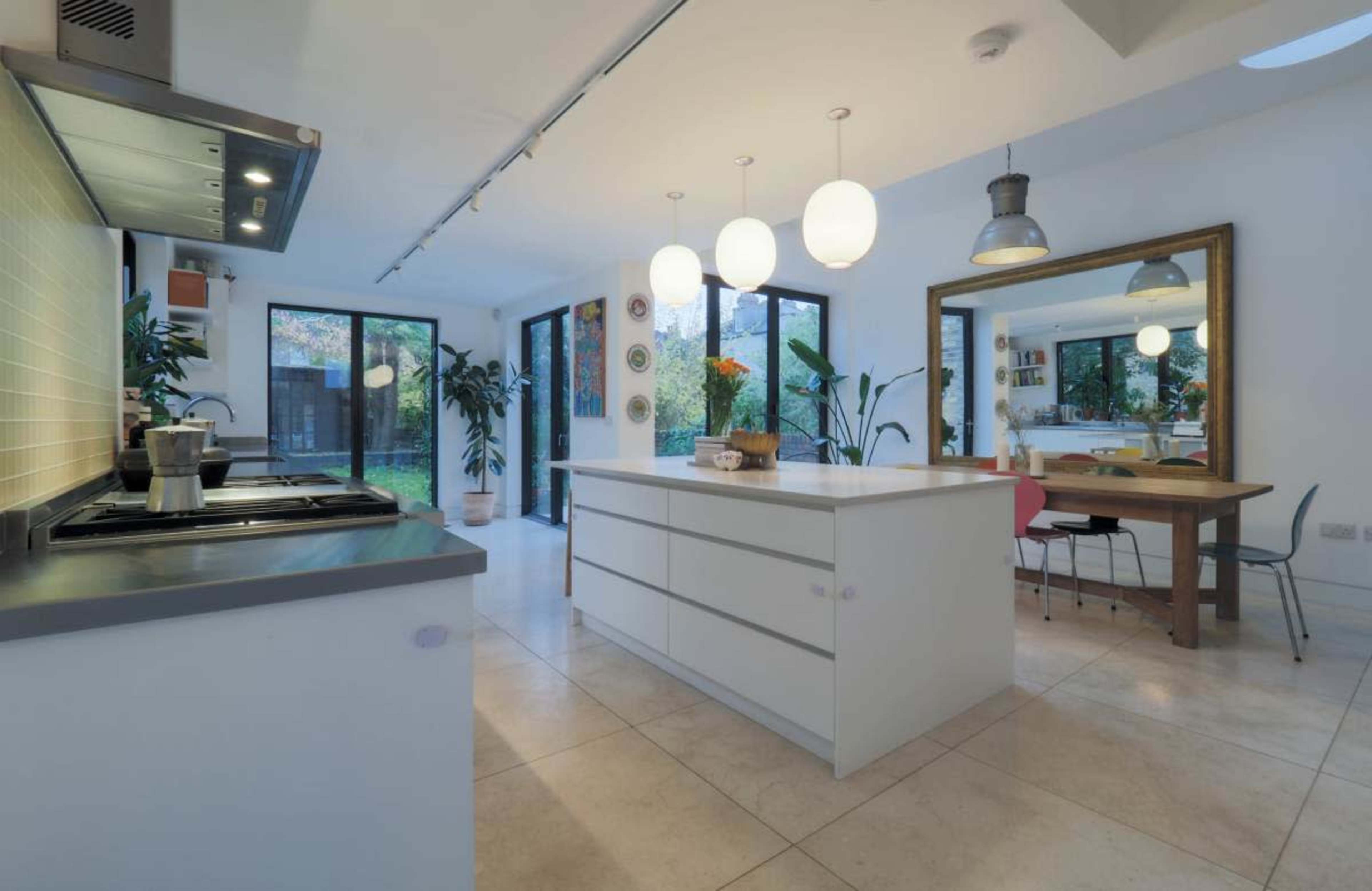 The image shows a modern kitchen with an island, featuring pendant lights, large windows, and a dining area reflected in a nearby mirror.