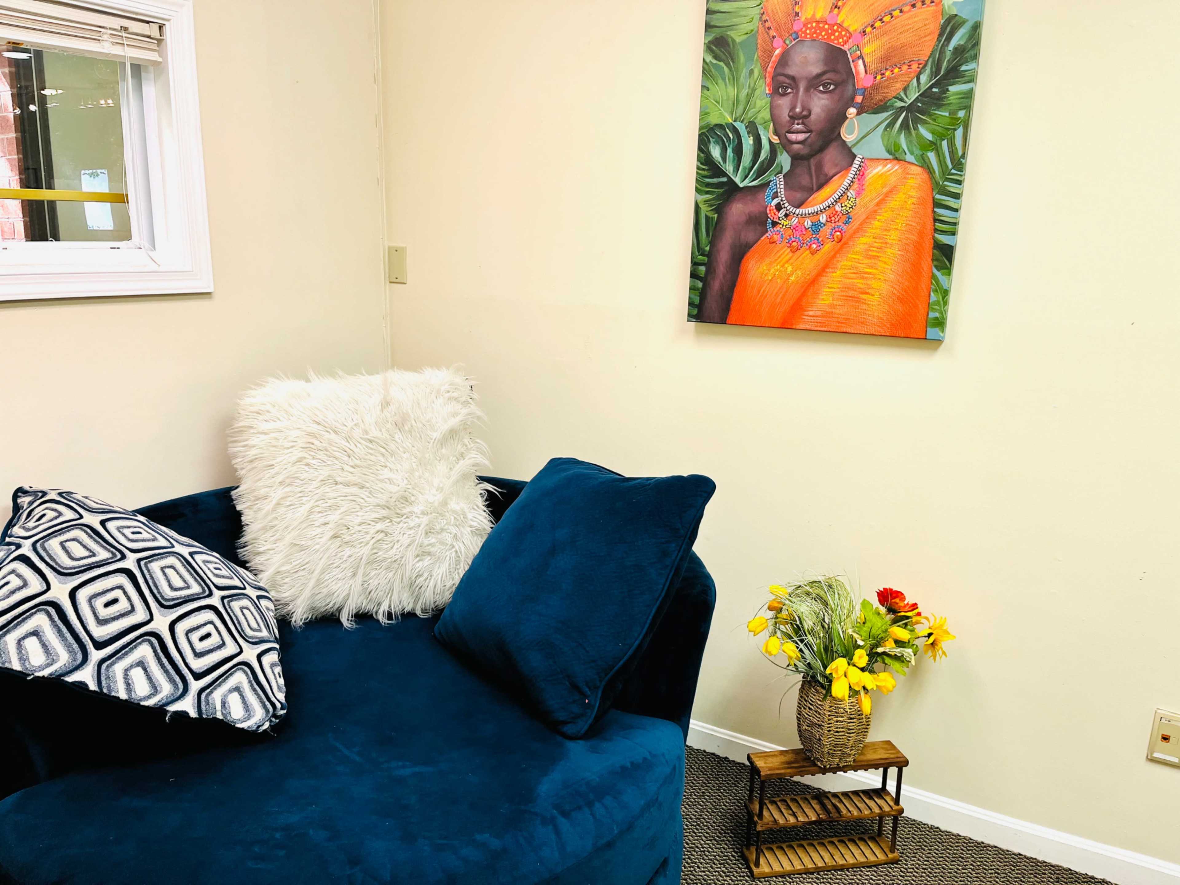 A blue upholstered chair with decorative pillows sits beside a small wooden stand holding a vase of flowers, while a colorful portrait hangs on the wall.