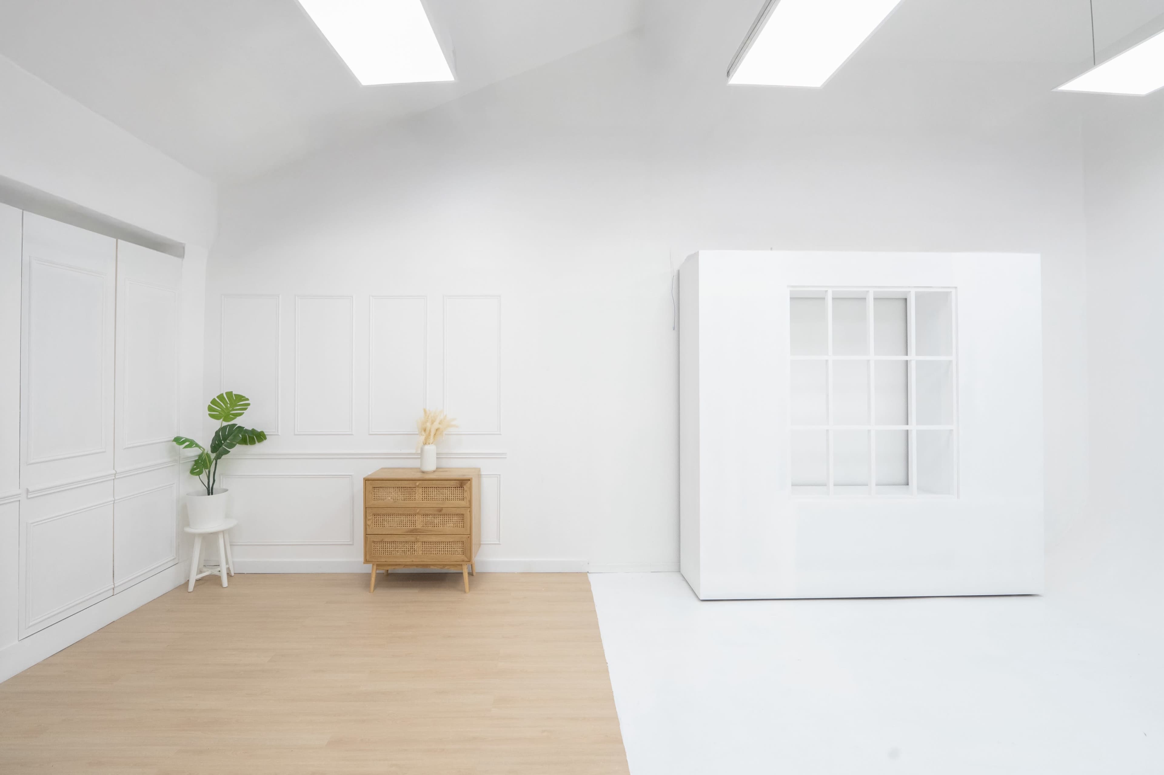 The image shows a bright, minimalist room with white walls, a light wooden floor, a wooden dresser, a small chair, and a large white display box with windows.