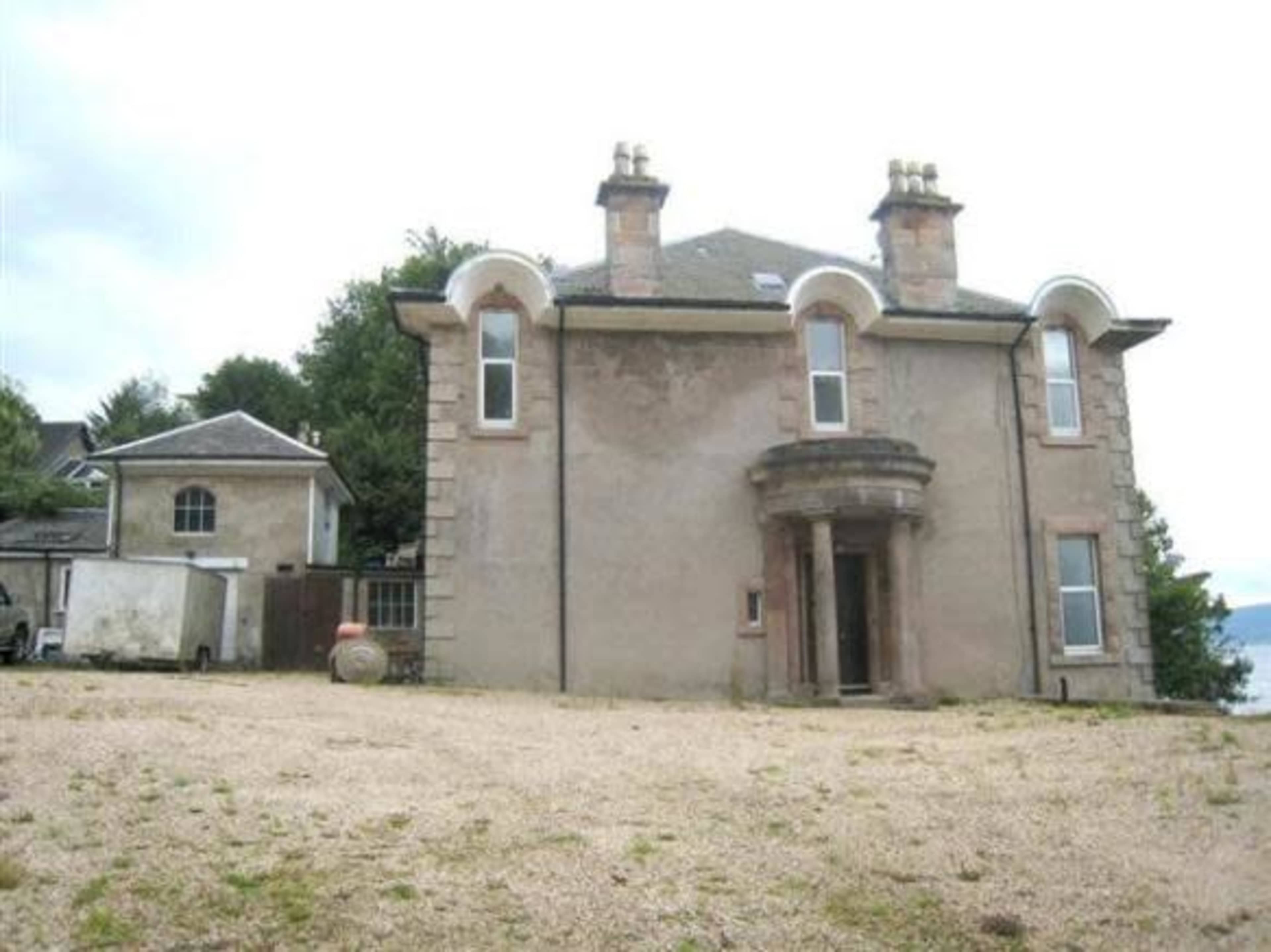 A large, old mansion with a central entrance and a gravel area in front, surrounded by trees and a nearby outbuilding.