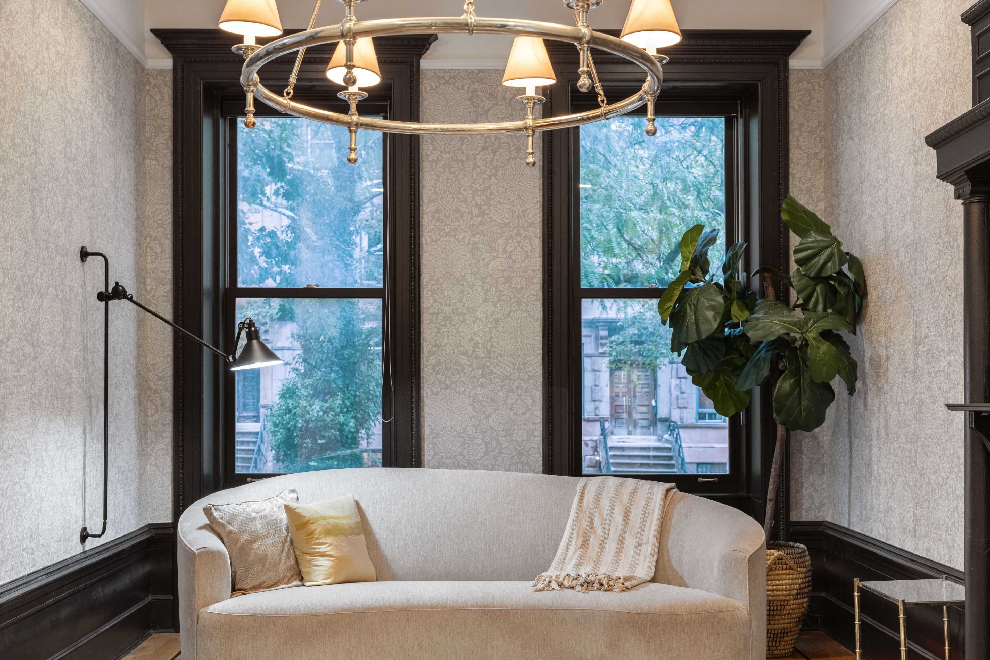 A light-colored sofa is positioned in a room with large windows, a modern chandelier, and a potted plant in the corner.