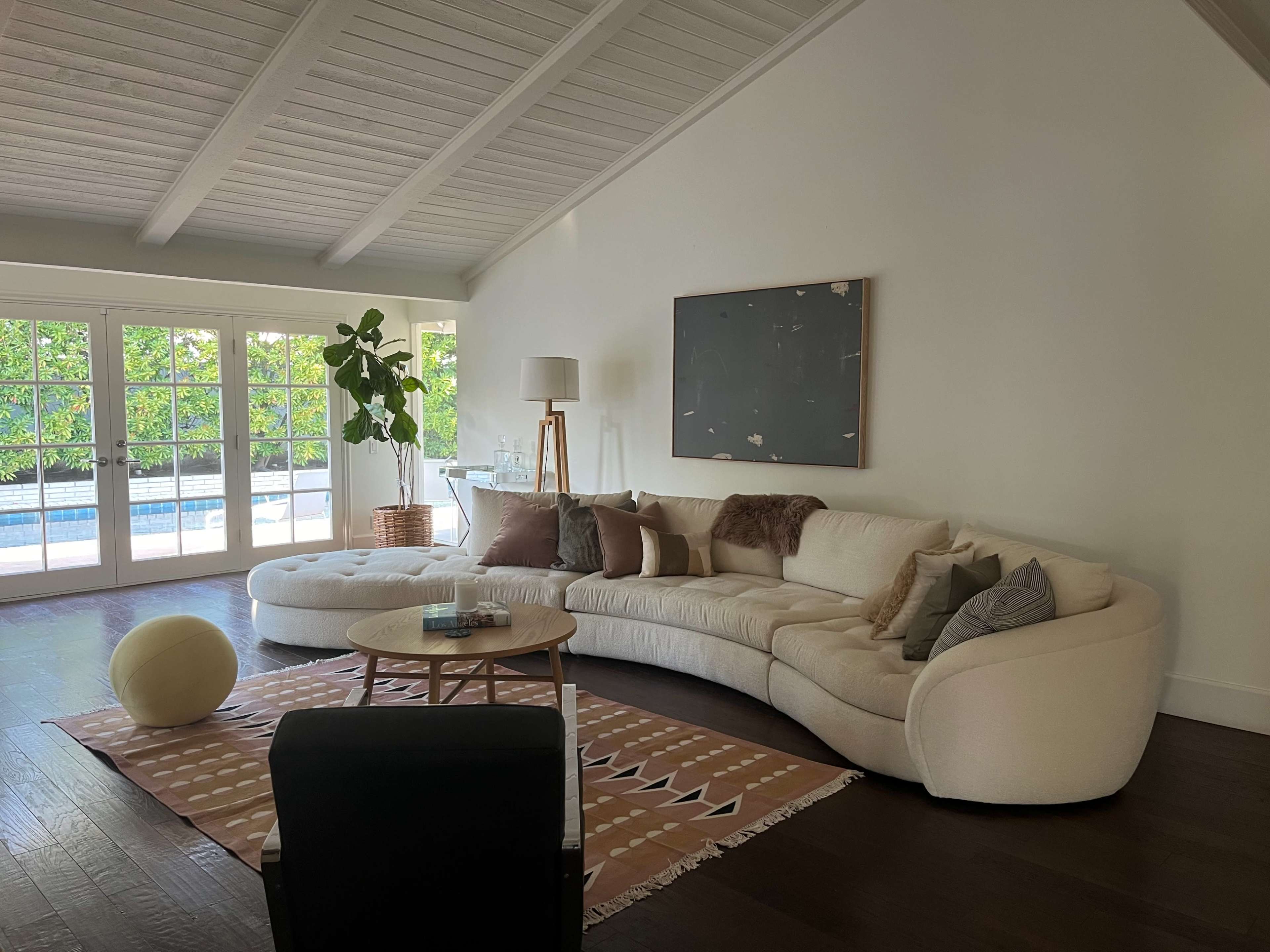 The image shows a modern living room featuring a curved sofa, a circular coffee table, and large windows that open to a pool area.