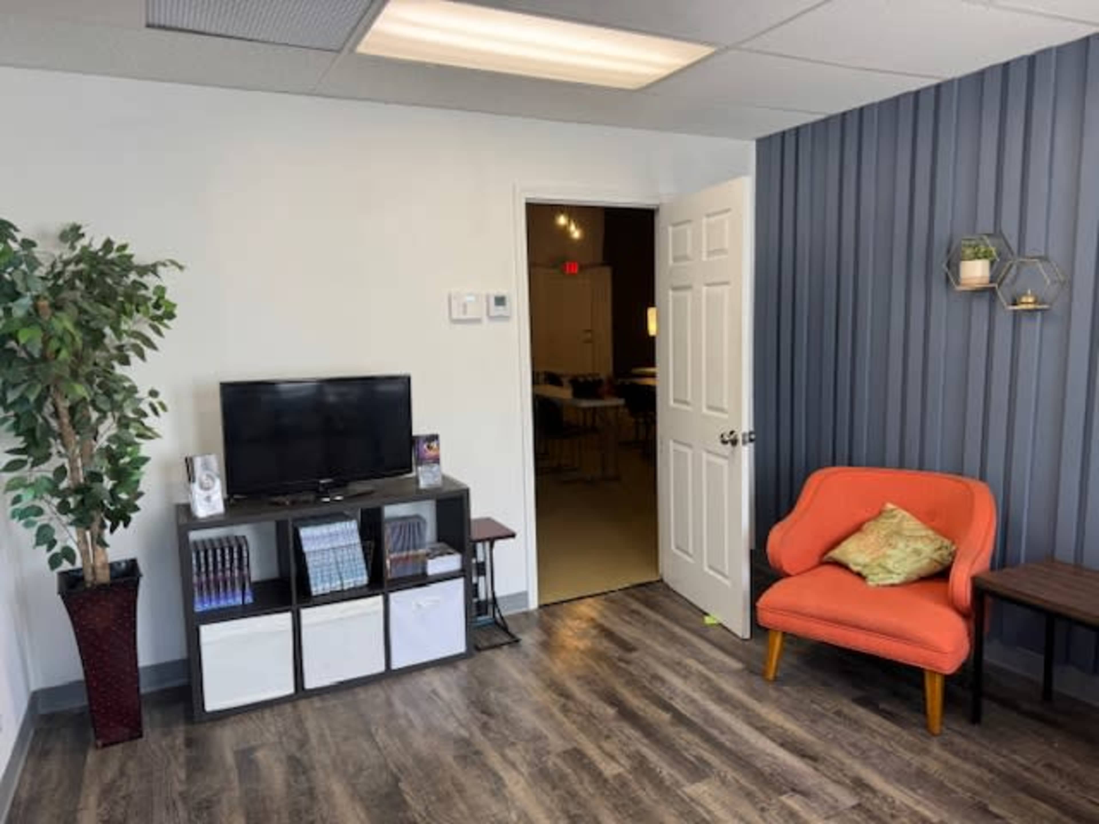 The image shows a small office or waiting area with a television, a potted plant, an orange armchair, and a door leading to another room.