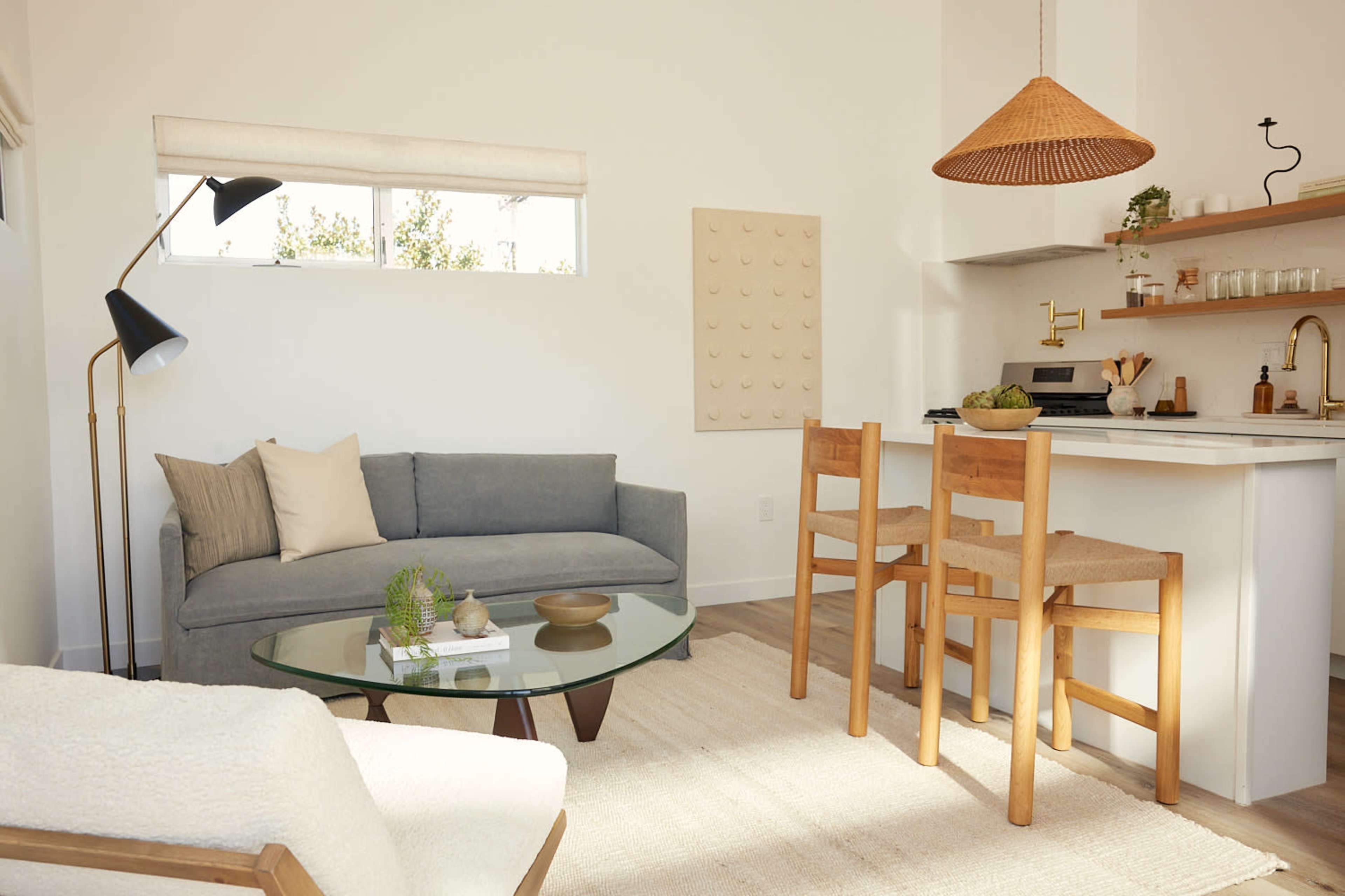 A modern living space features a gray sofa, a glass coffee table, a white dining counter with two wooden chairs, and natural light coming through the windows.