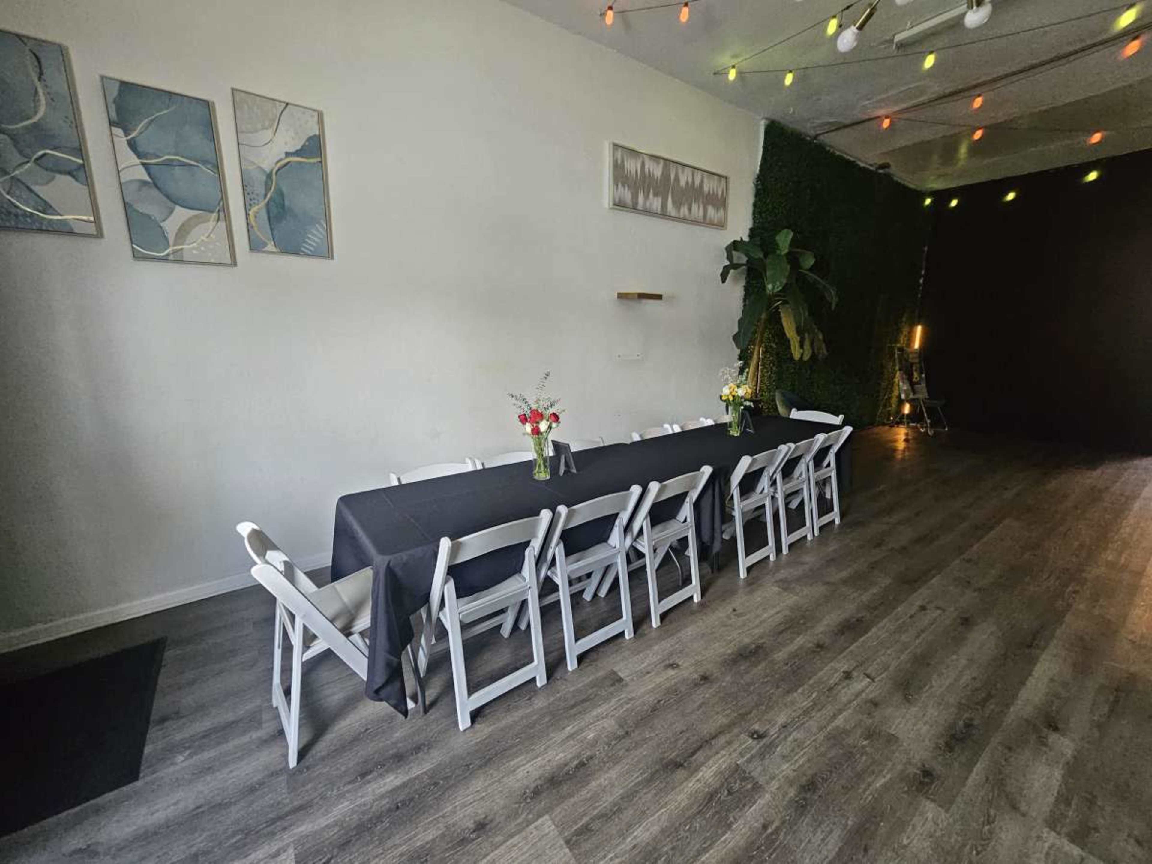 A long table set with a black cloth and white chairs is positioned in a spacious, well-lit room featuring greenery along the walls and colorful string lights.