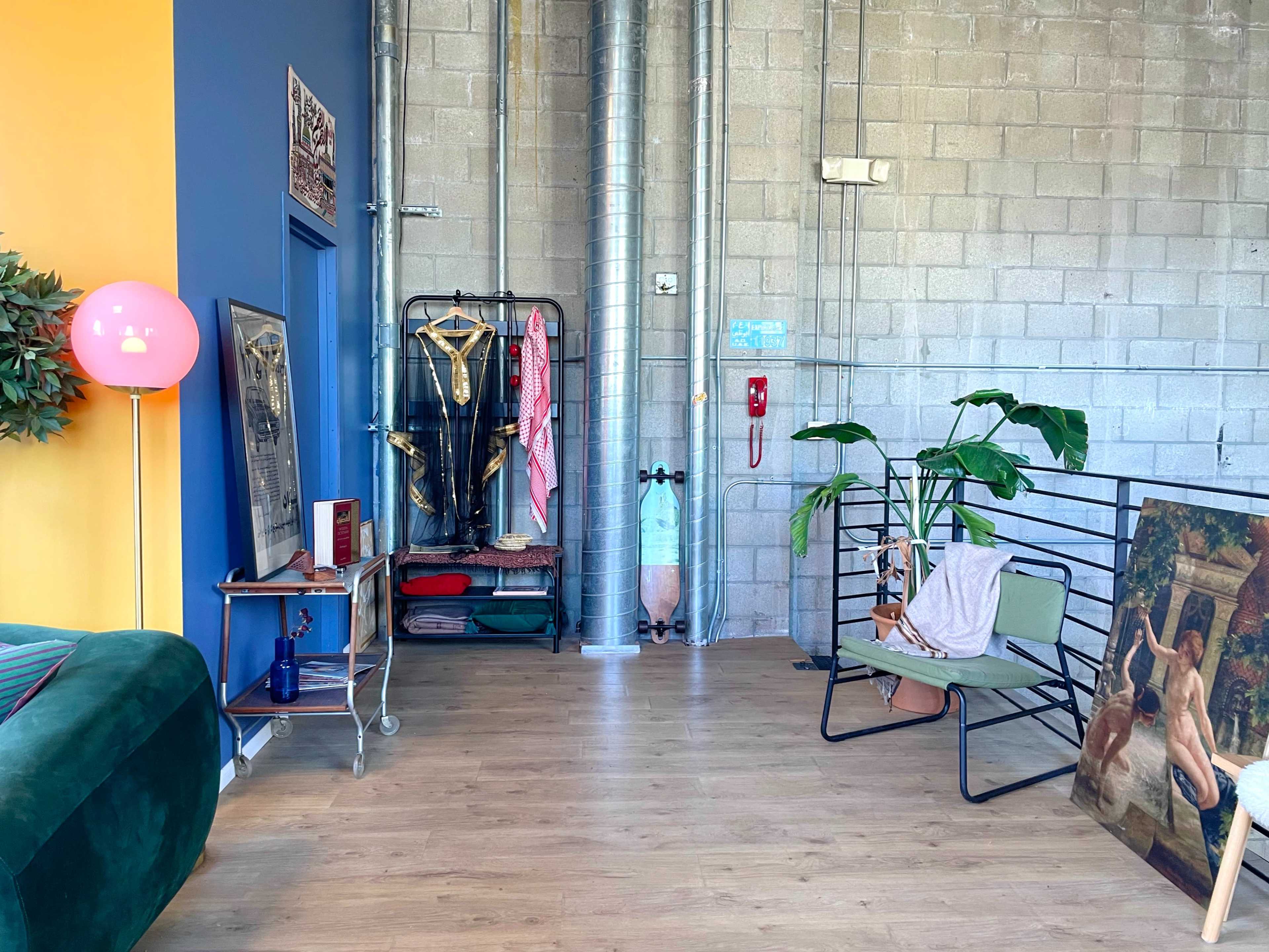 The image shows a modern industrial-style room with a green sofa, a pink floor lamp, a clothing rack, a potted plant, and various artworks leaning against the walls.