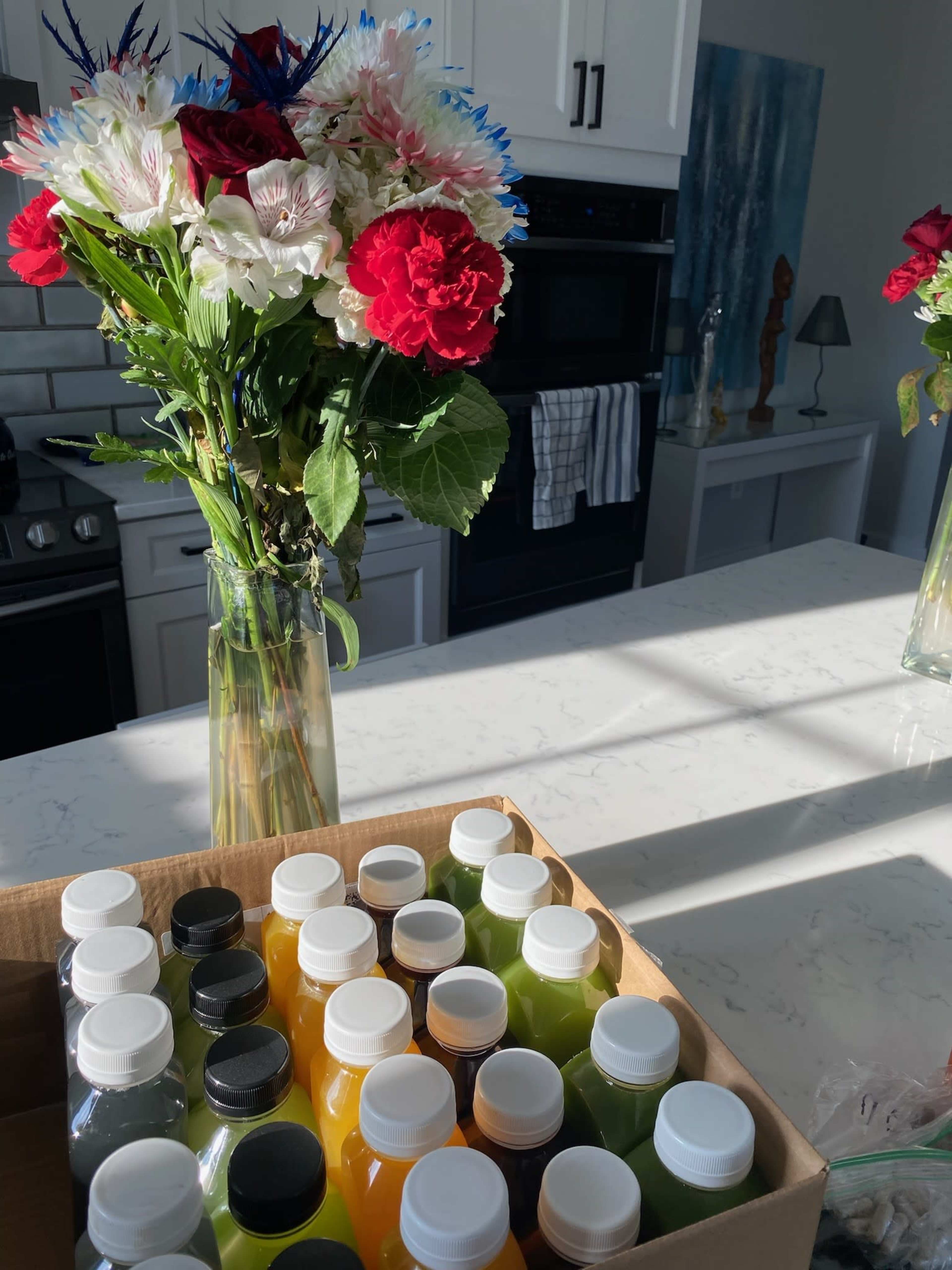 A bouquet of flowers sits in a vase on a kitchen counter next to a box of colorful bottled drinks.