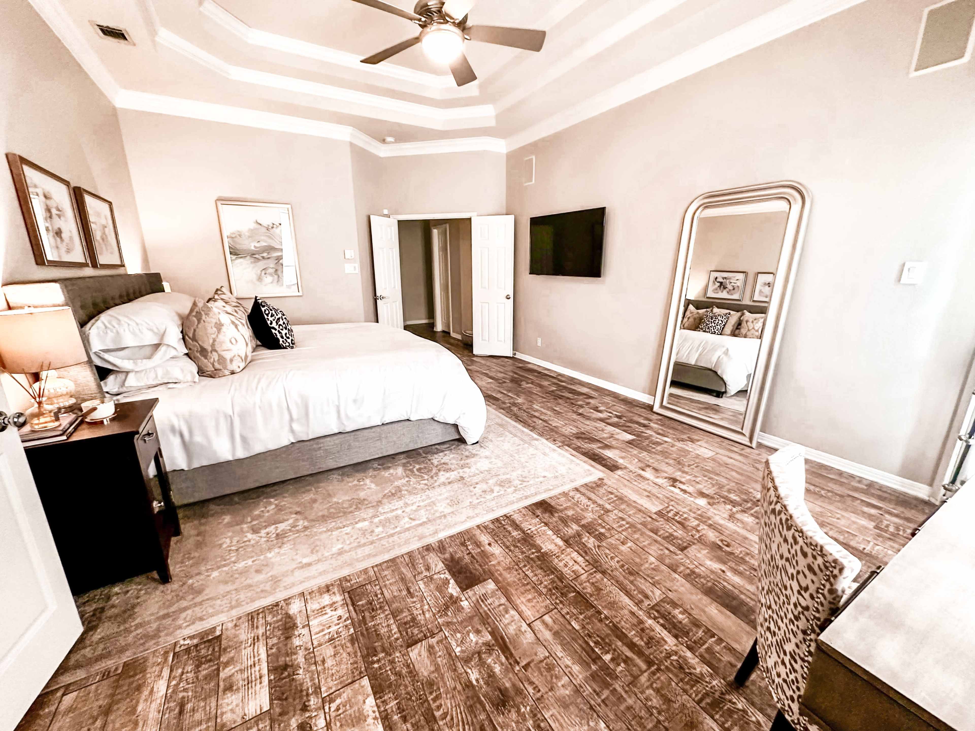 The image shows a spacious bedroom featuring a large bed, a desk chair, a wall-mounted television, and a wall mirror, with hardwood flooring and neutral-colored walls.