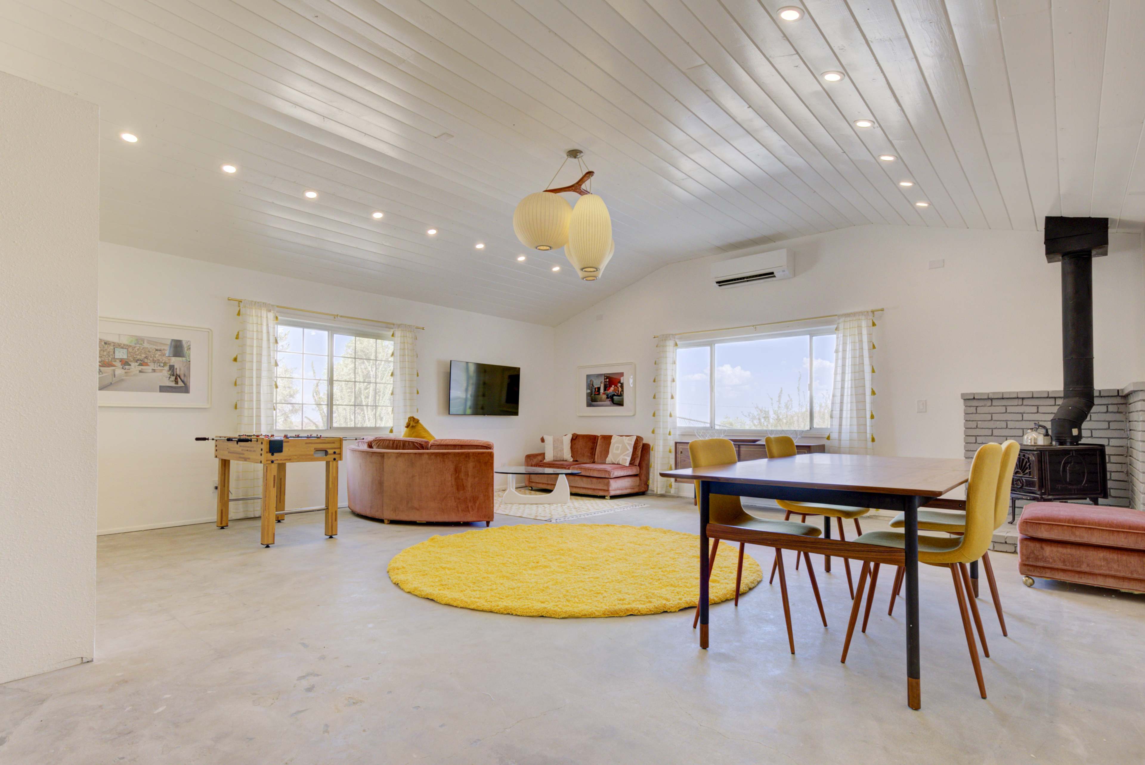 The image shows a spacious living area with a yellow rug, a dining table, a foosball table, and a seating arrangement, featuring a wood-burning stove and large windows.