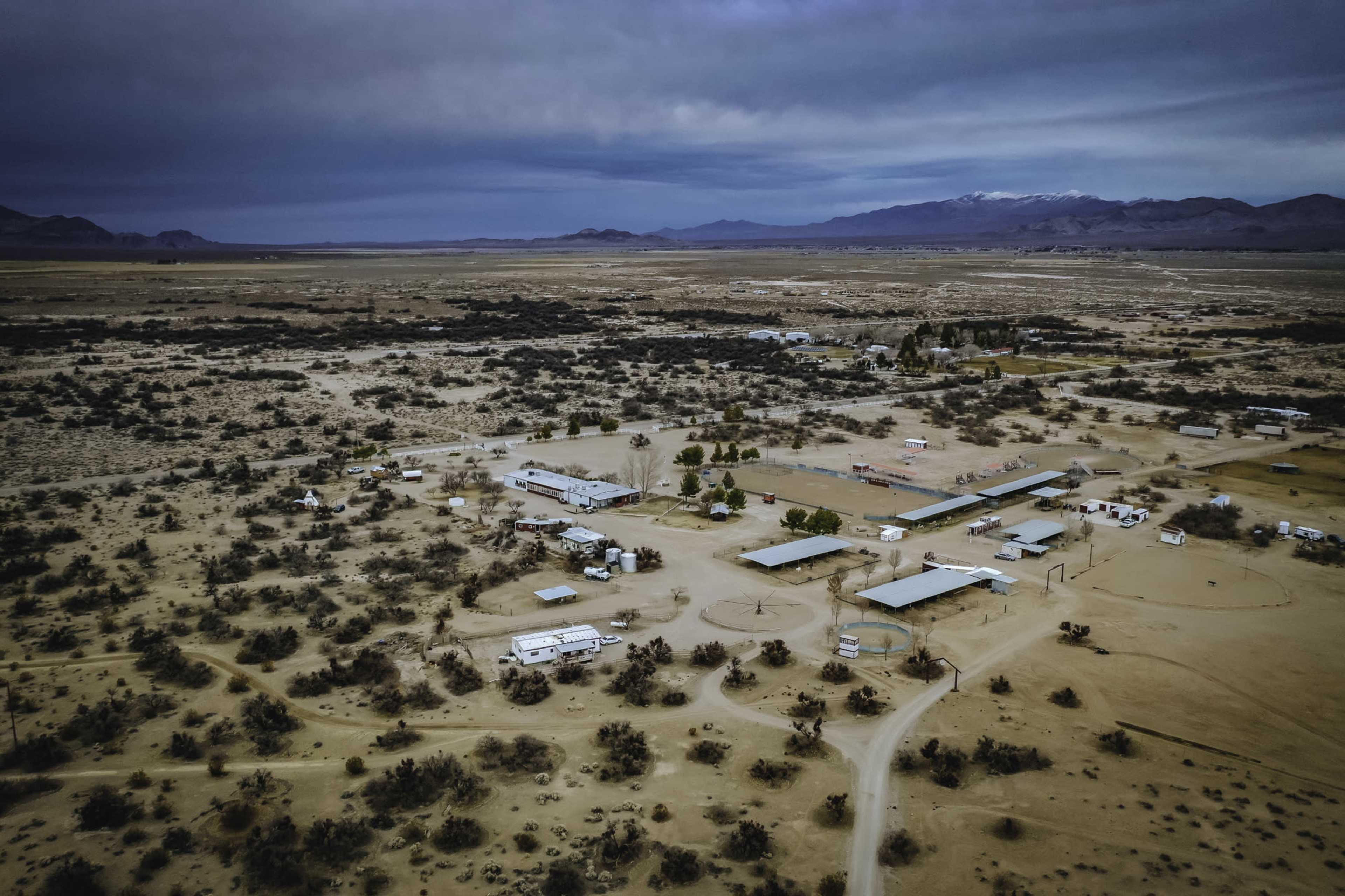 An aerial view shows a small settlement surrounded by desert terrain and sparse vegetation, featuring several buildings and roads.