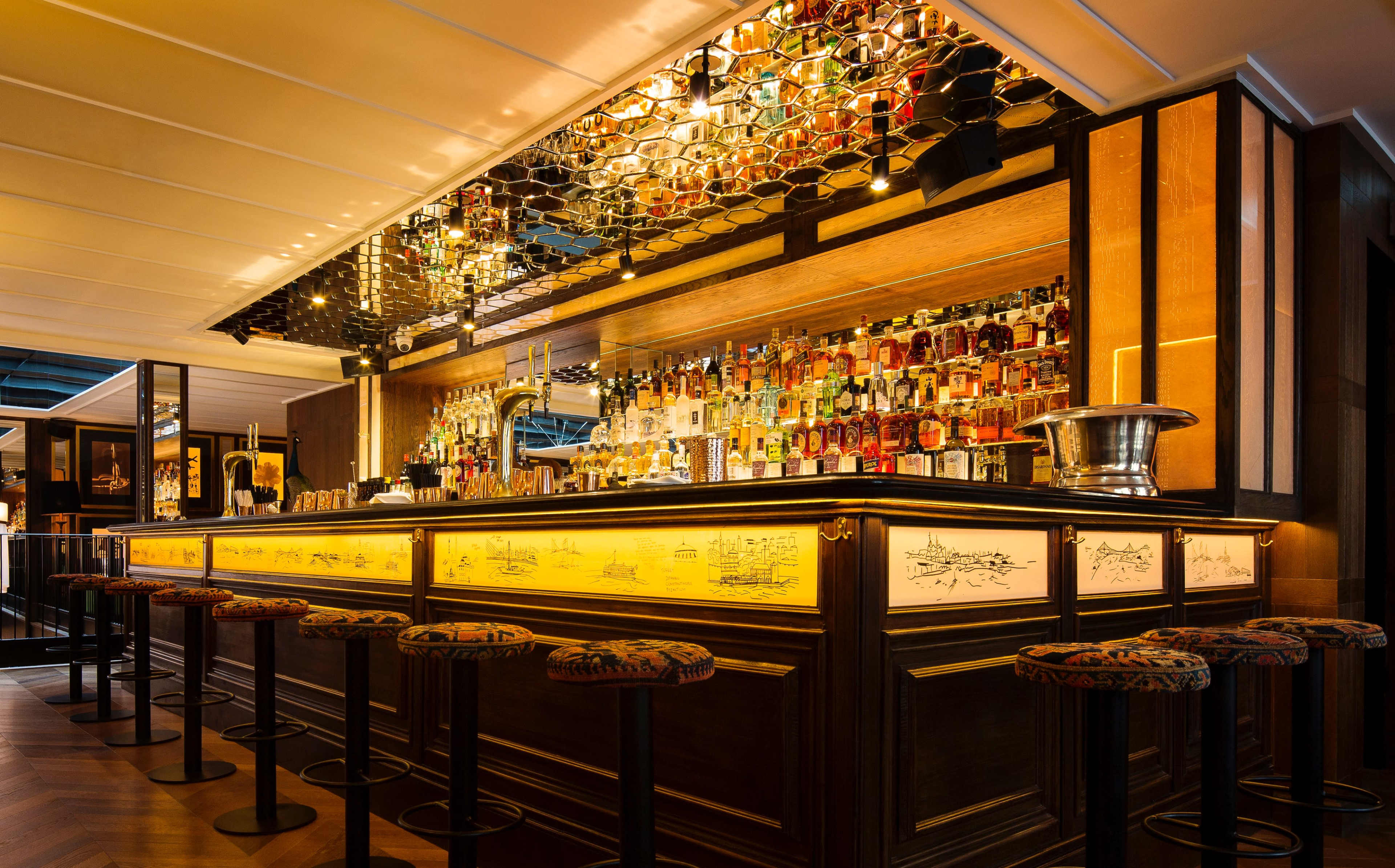 The image shows a stylish bar with a brightly lit backlit shelf displaying various bottles, complemented by elegant bar stools.