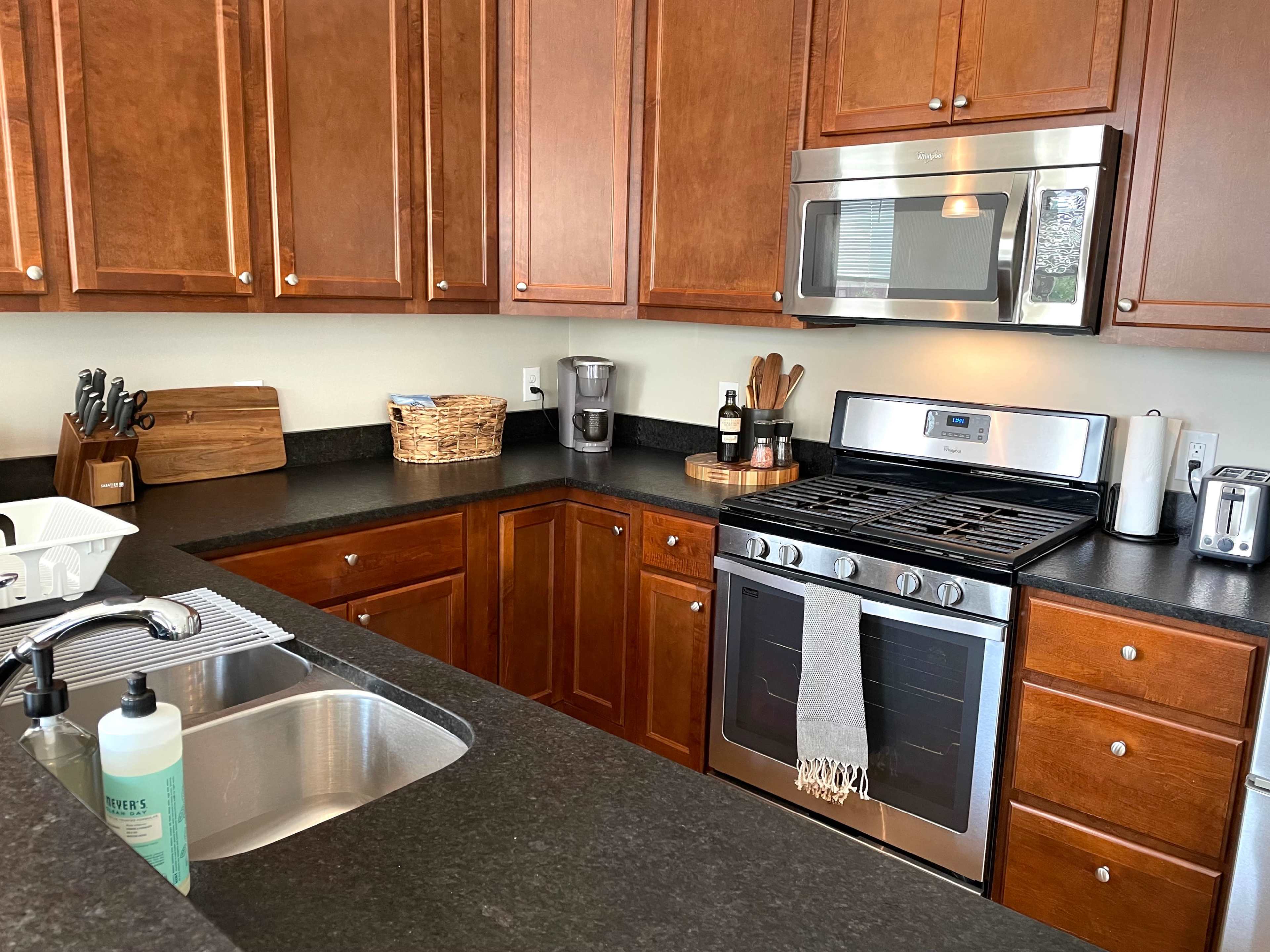 A modern kitchen features wooden cabinets, stainless steel appliances, and a black countertop with a sink and various utensils.