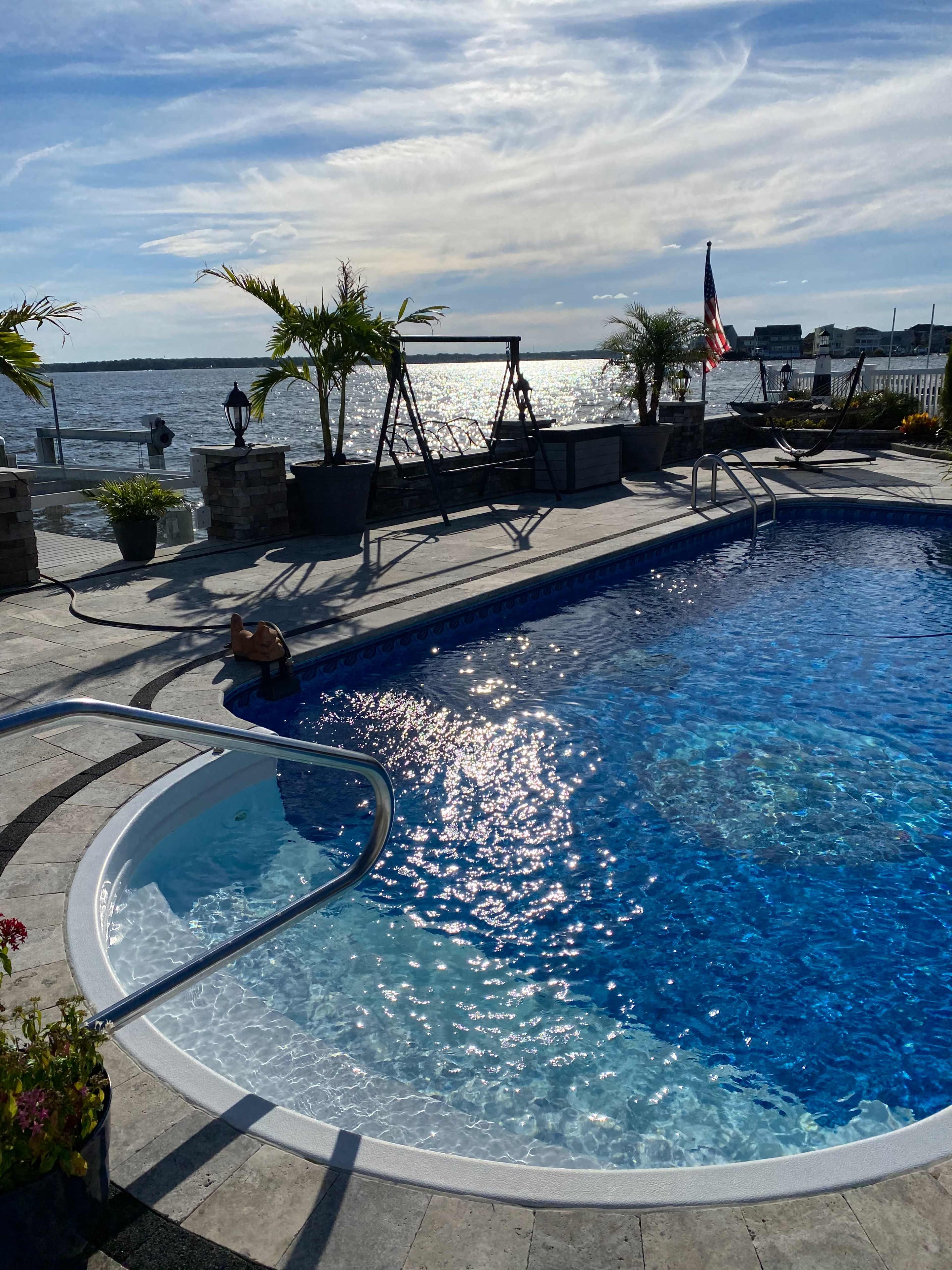 The image shows a swimming pool with clear blue water, surrounded by palm trees and a view of the water in the background under a partly cloudy sky.