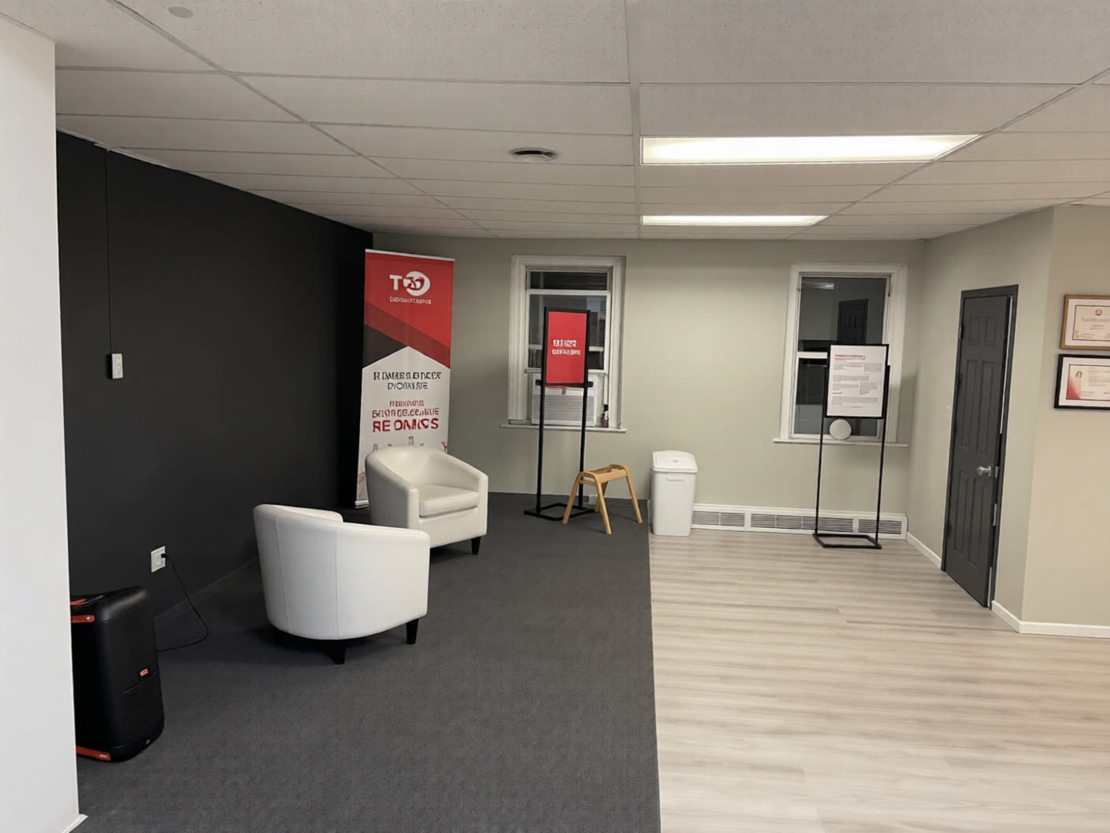 The image shows a modern office space with two white chairs, a black wall, a banner, and a waste bin in the corner.