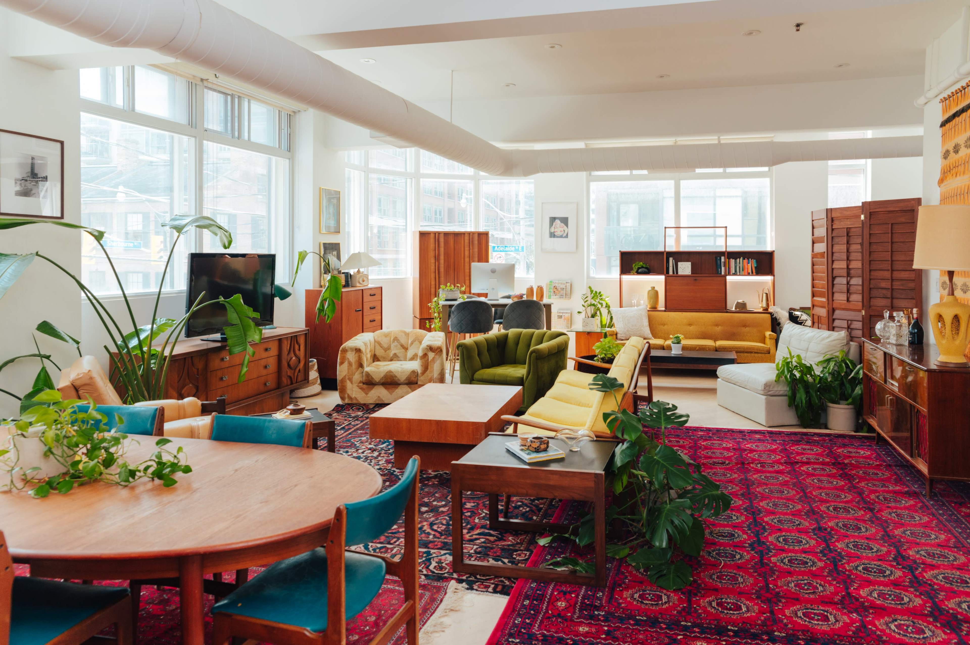 A spacious, well-lit living area featuring various furniture pieces, including a large dining table, several couches, and potted plants on a patterned rug.
