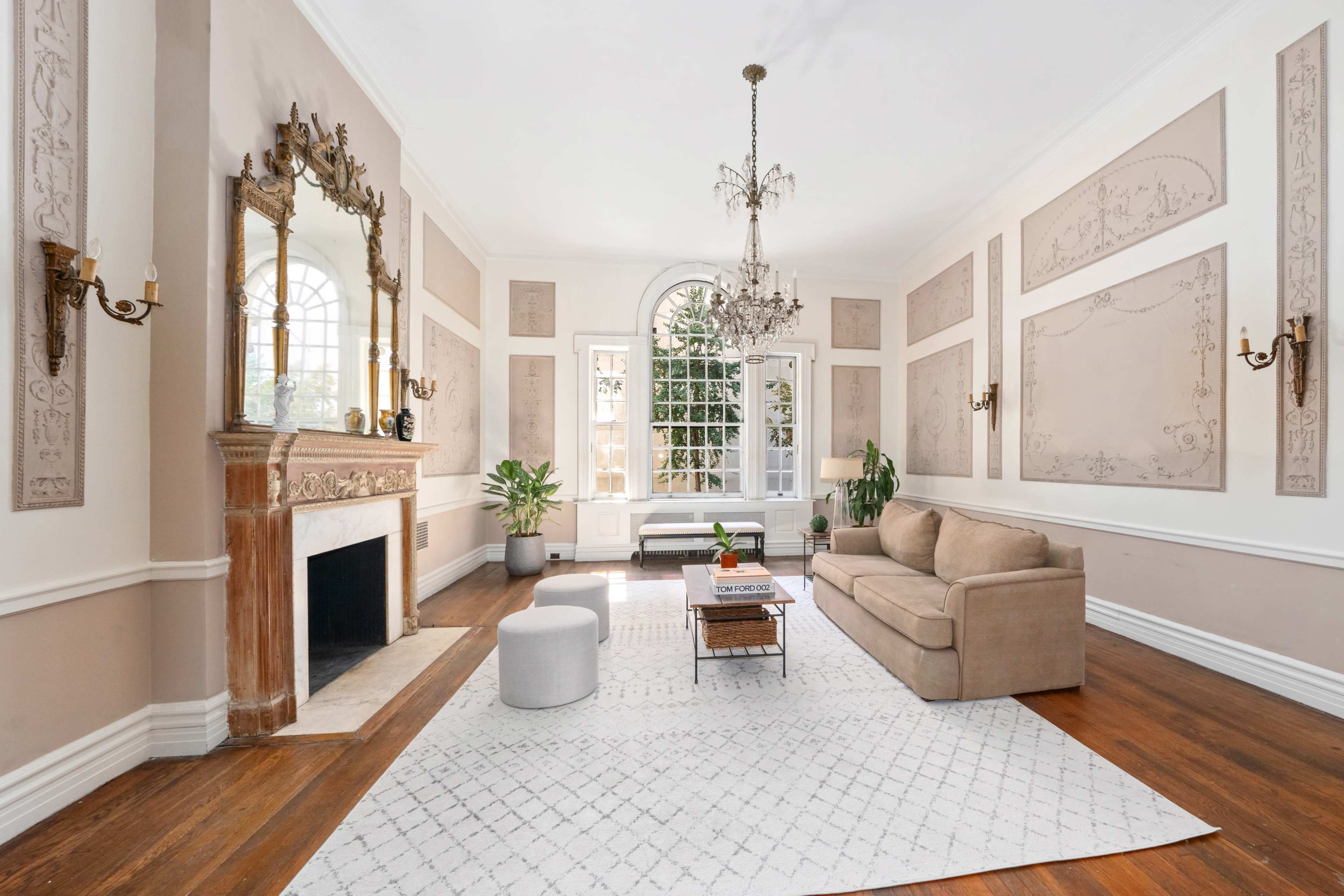 A spacious living room with a beige sofa, a patterned area rug, a marble fireplace, and large windows allowing natural light.