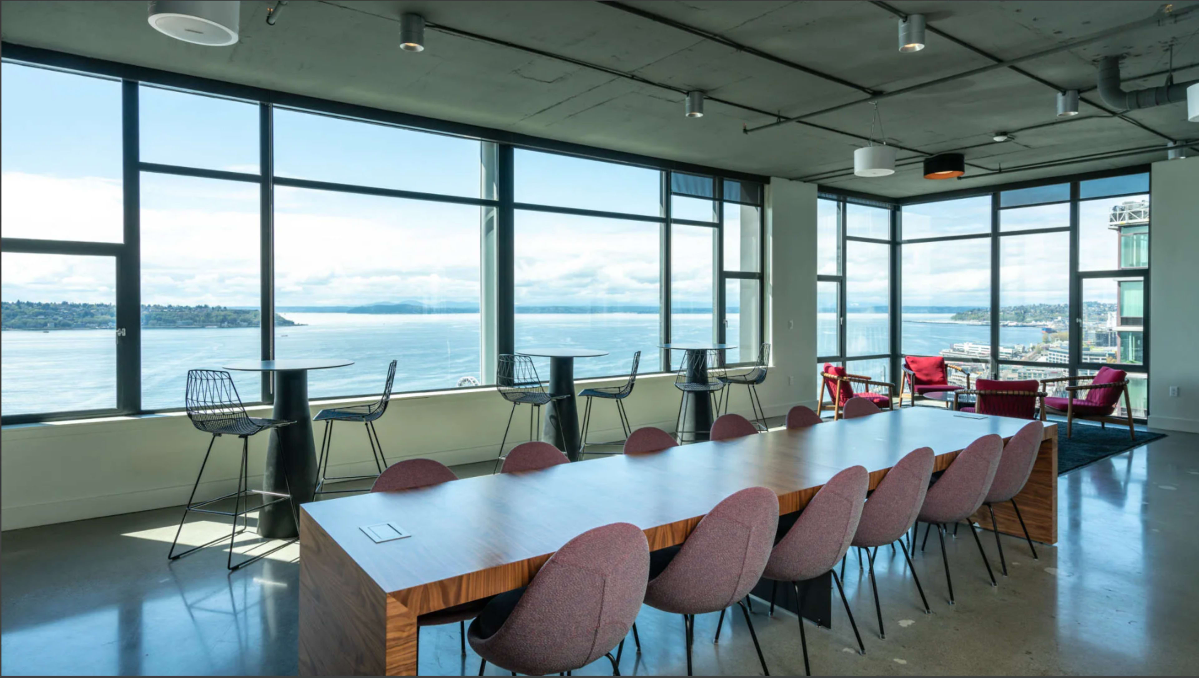 A modern conference room with a long wooden table and eight patterned chairs, featuring large windows that offer a view of the water and cloudy sky outside.