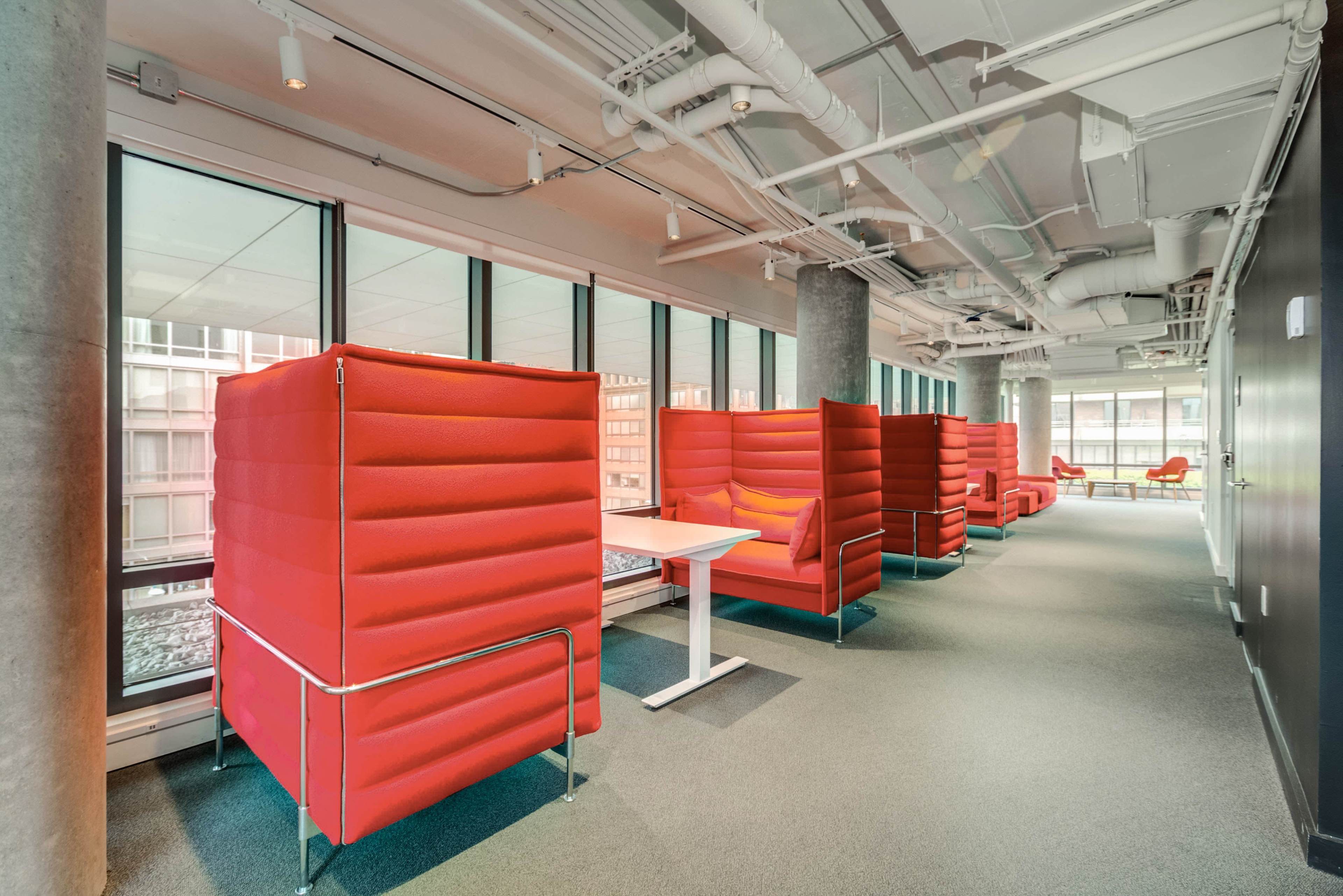 The image shows a modern office space featuring red booth seating arranged along large windows, with a table in the center for collaborative work.