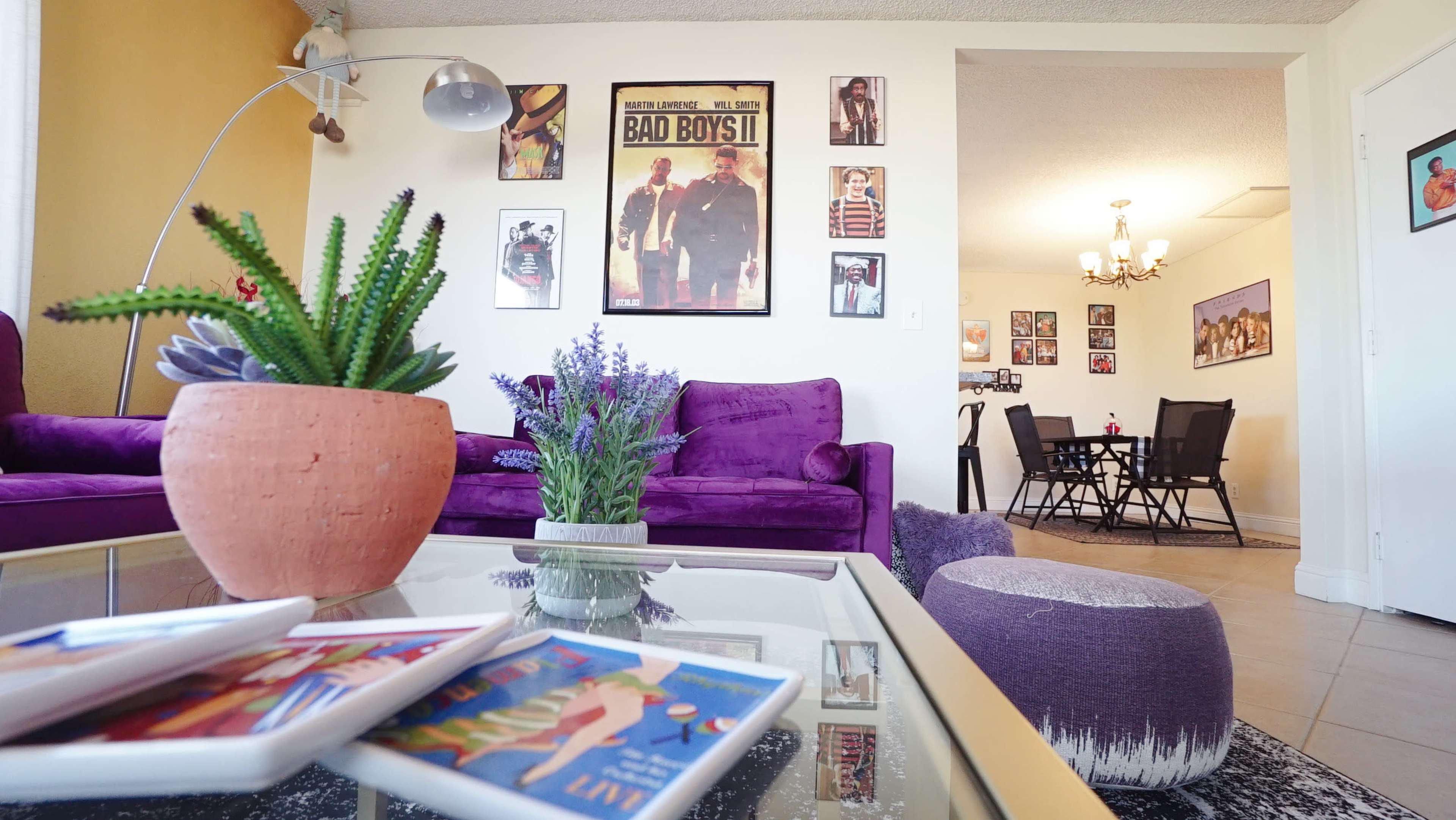 A living room features a purple sofa, a potted plant, a coffee table with decorative plates, and wall art, with a dining area visible in the background.