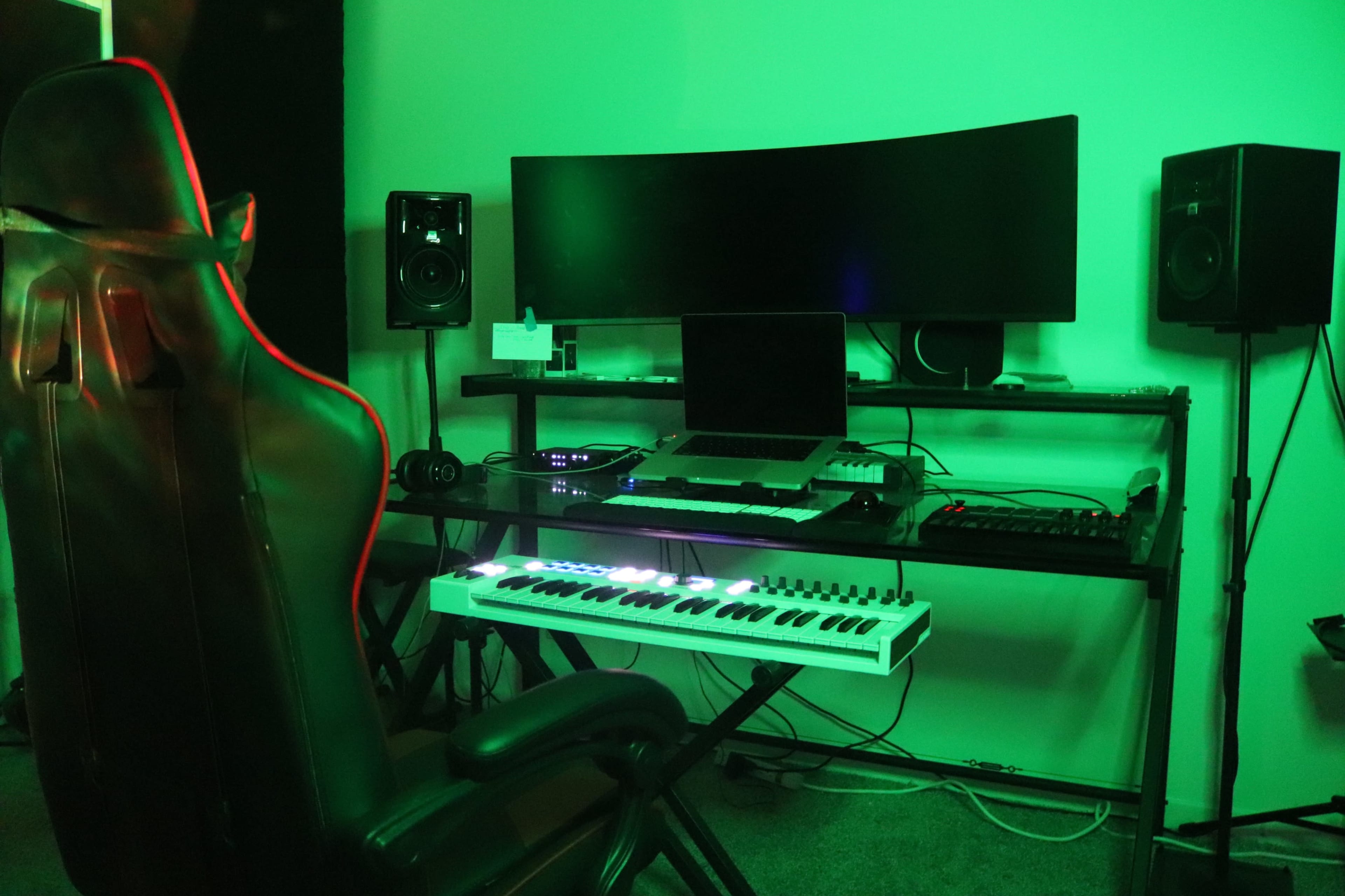 A music production setup features a curved monitor, various equipment, and a keyboard, all illuminated by green lighting.