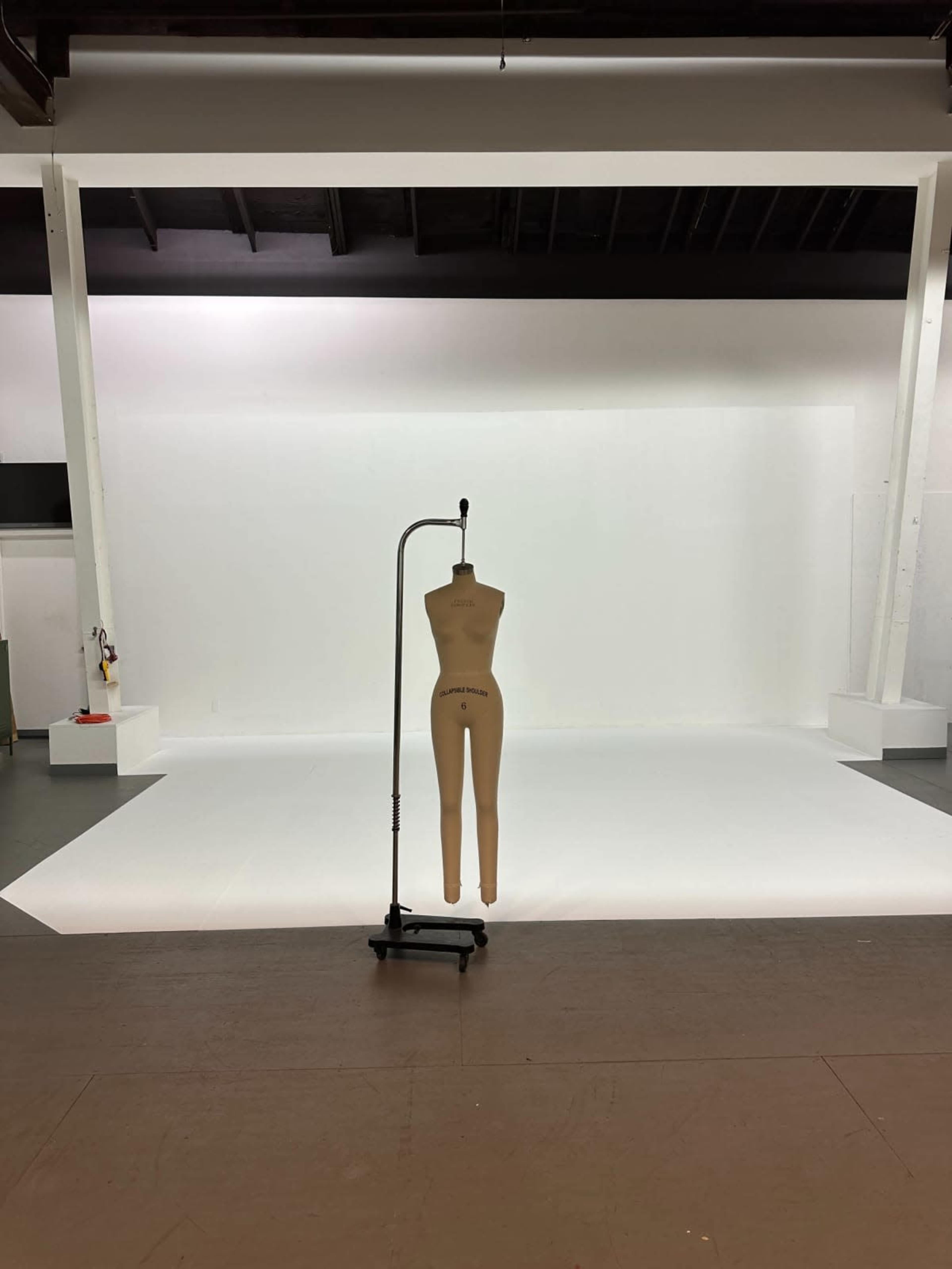 A dress form is positioned on a stand in front of a blank white backdrop in a studio space.
