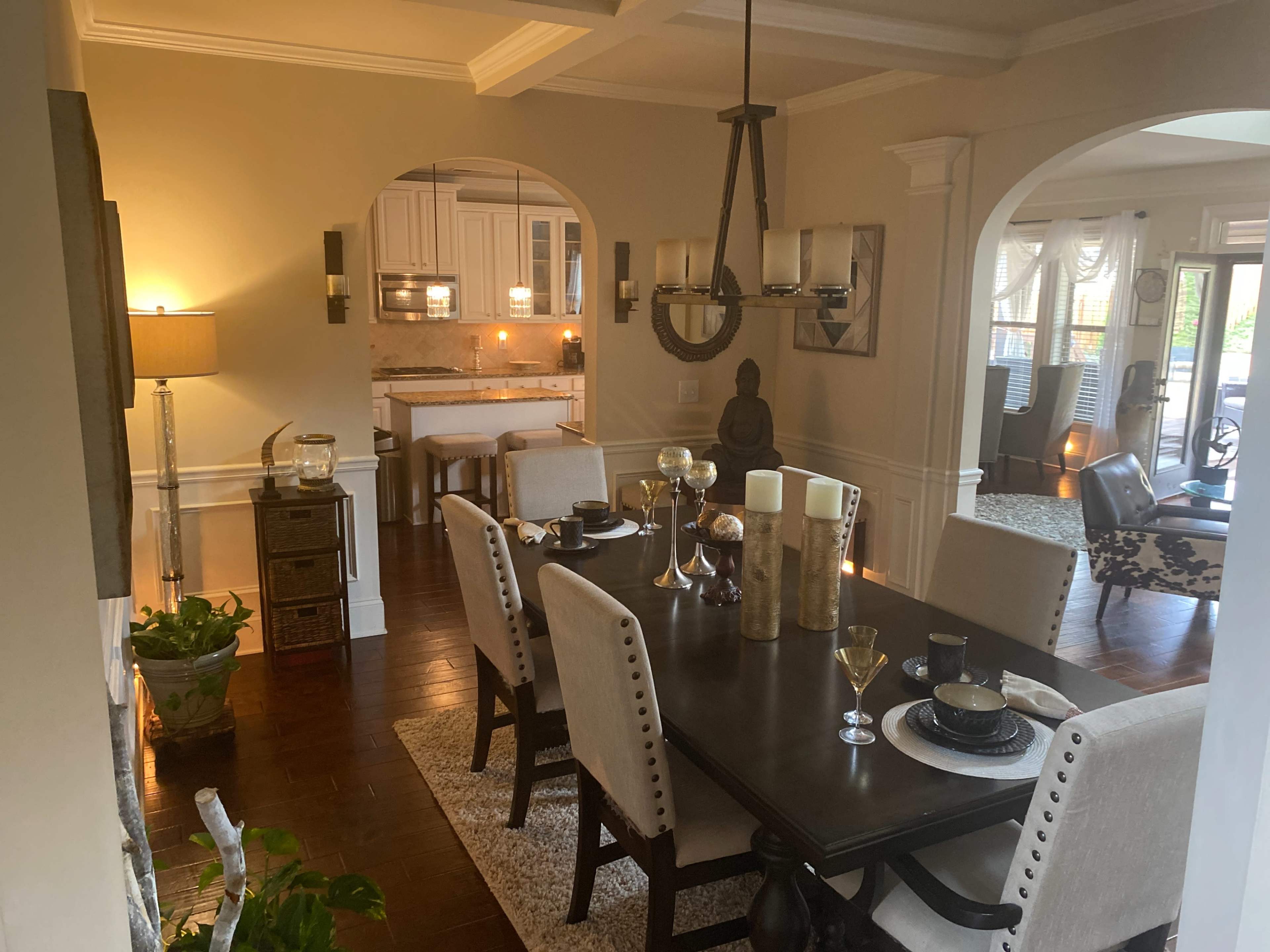 A dining area with a wooden table set for a meal connects to a kitchen through an arched entryway, while the room is illuminated by light fixtures and natural light.