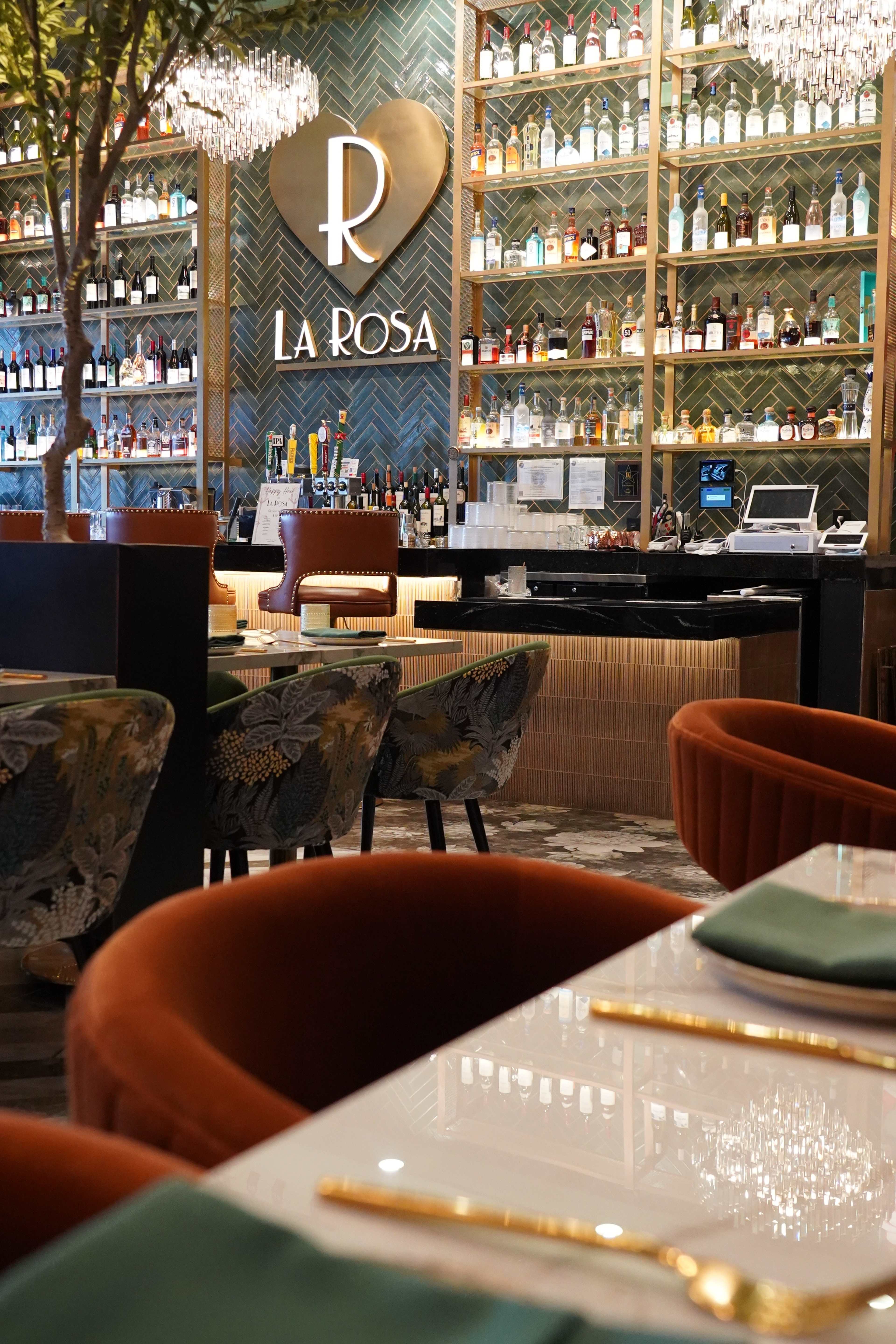 The image shows an elegant bar area at a restaurant called "La Rosa," featuring shelves stocked with various bottles of liquor, plush seating, and decorative lighting.